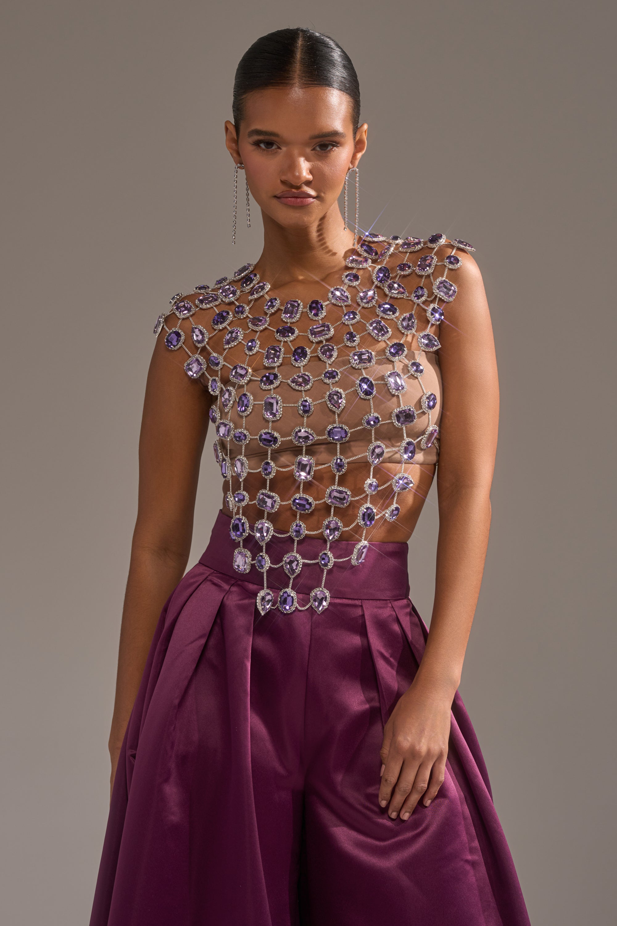 A woman wears high-waisted, deep purple wide-leg pants and the LOVER GIRL RHINESTONE CHAIN TOP adorned with large, sparkling purple gemstones, standing against a plain gray background. 4