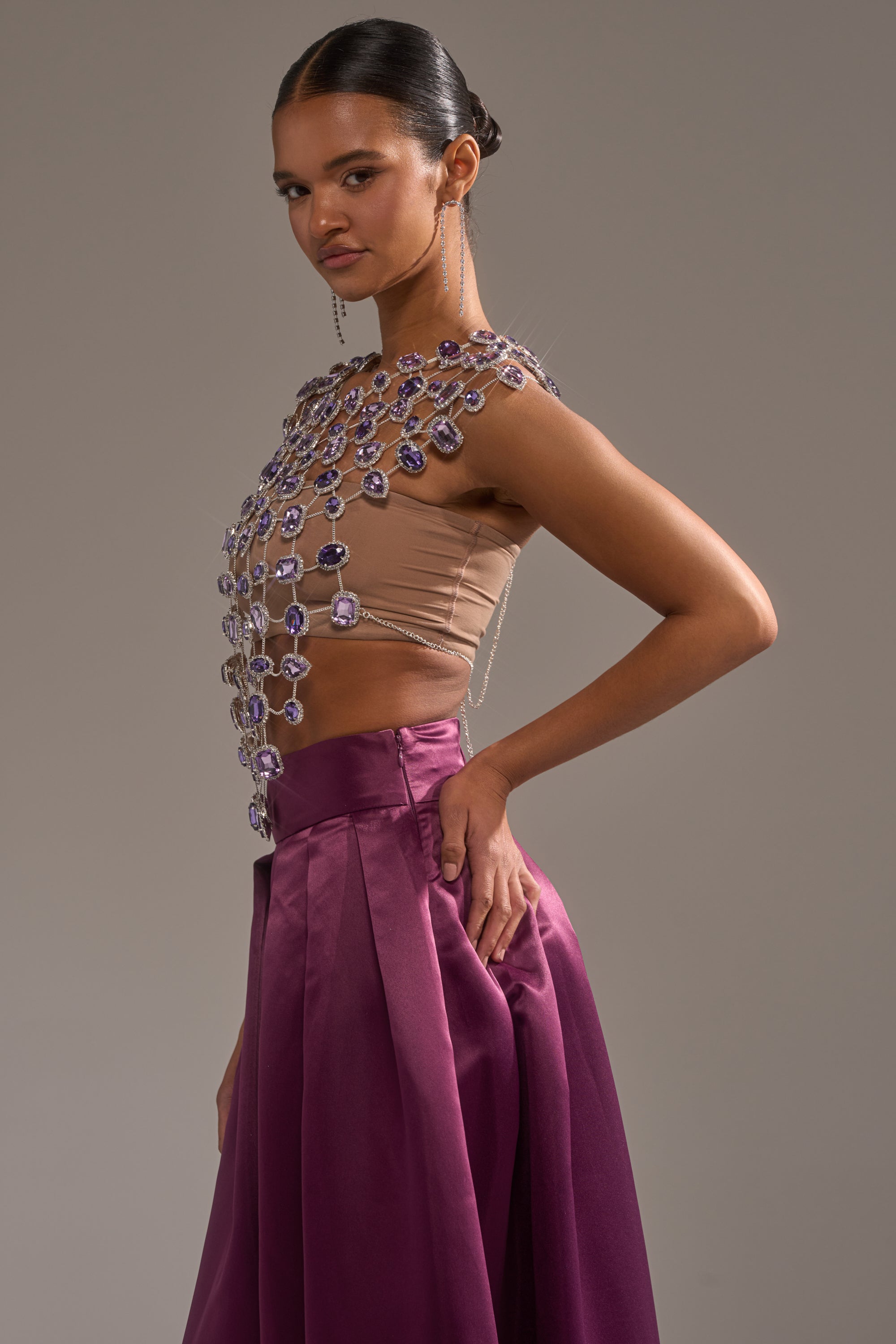 A woman poses in profile wearing the LOVER GIRL RHINESTONE CHAIN TOP with large purple gemstones and a high-waisted, pleated, deep purple satin skirt against a neutral background. 5
