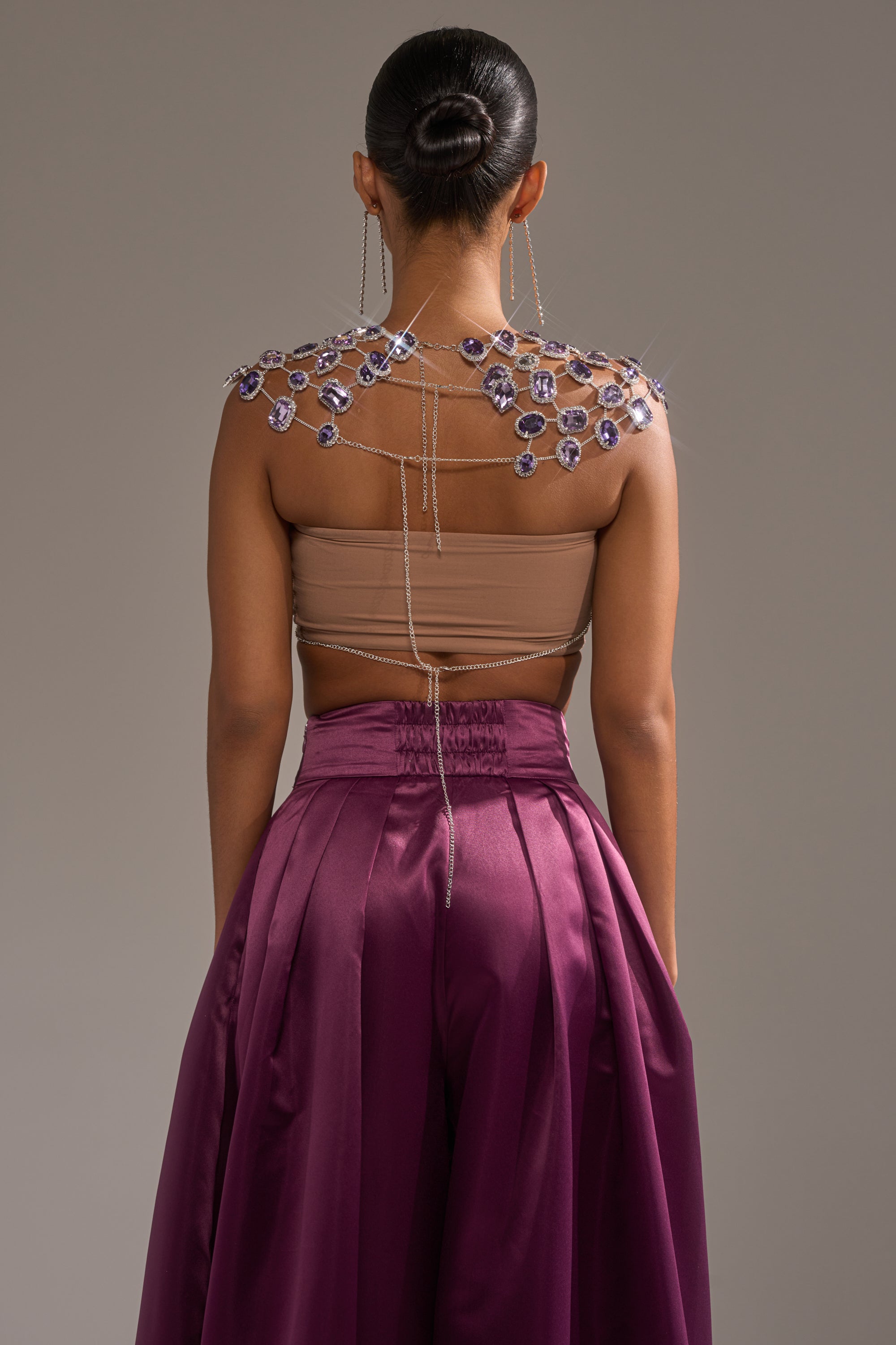 A woman with a sleek bun faces away from the camera, wearing the LOVER GIRL RHINESTONE CHAIN TOP with dazzling crystal shoulder chains and high-waisted, flowing purple satin pants. 6