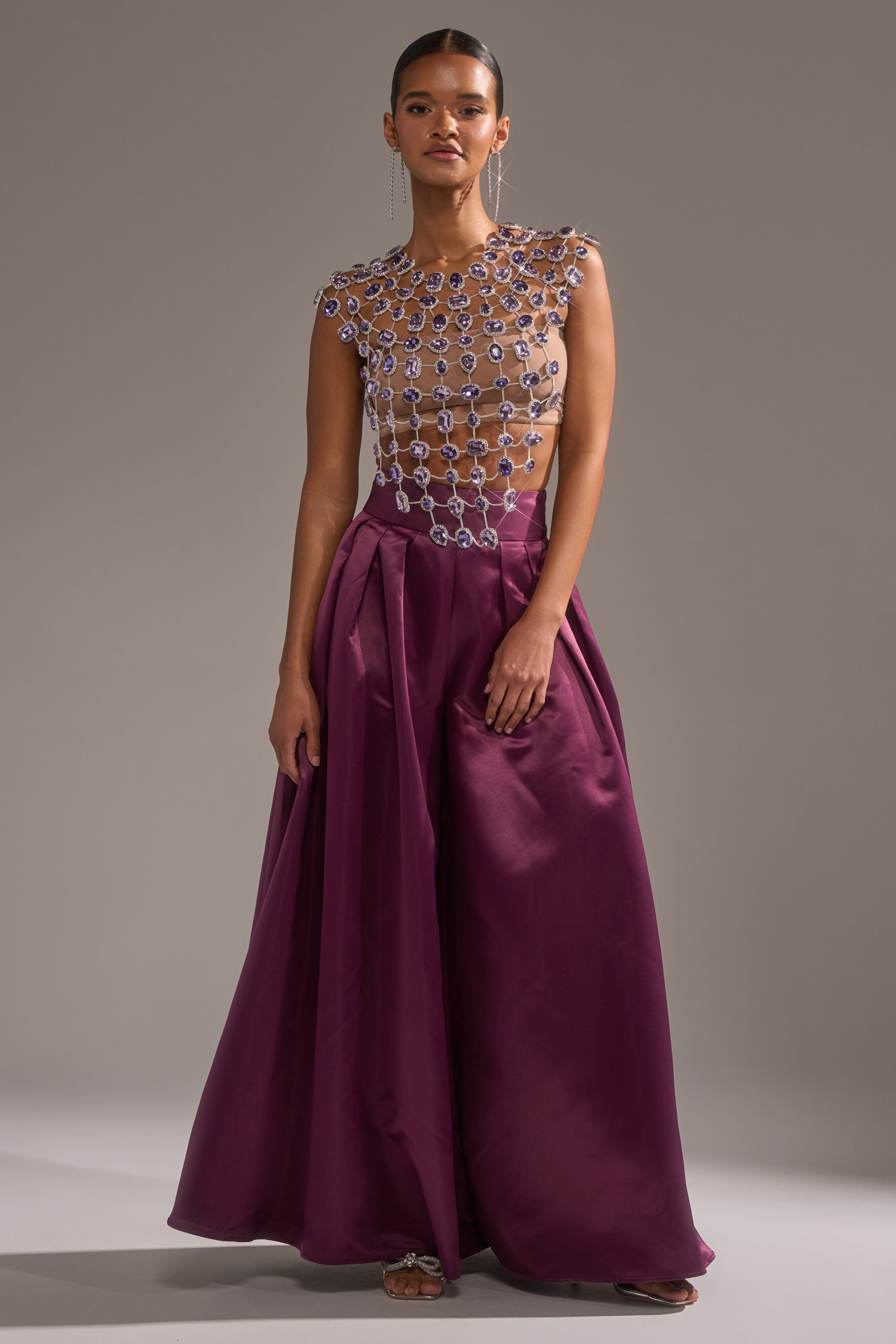 A woman exudes confidence in the LOVER GIRL RHINESTONE CHAIN TOP paired with a high-waisted, floor-length deep purple satin skirt, creating a striking silhouette against a plain gray background. 7