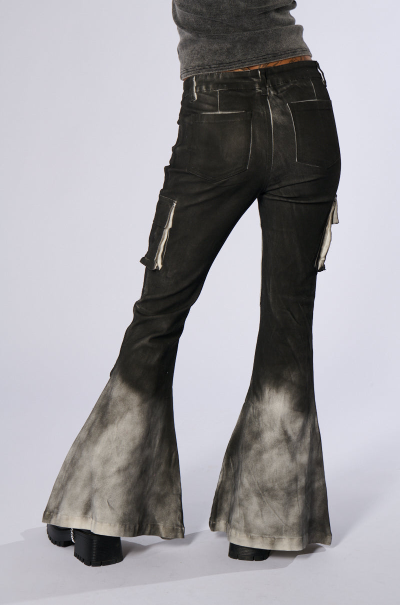 A person wearing the ROCK YOU ALL NIGHT LONG DENIM PANT, black high-waisted flare jeans featuring an artsy airbrushed design at the bottom, stands with their back to the camera. The jeans have white accents on the pockets and legs. They pair these stylish jeans with a grey top and black platform shoes.