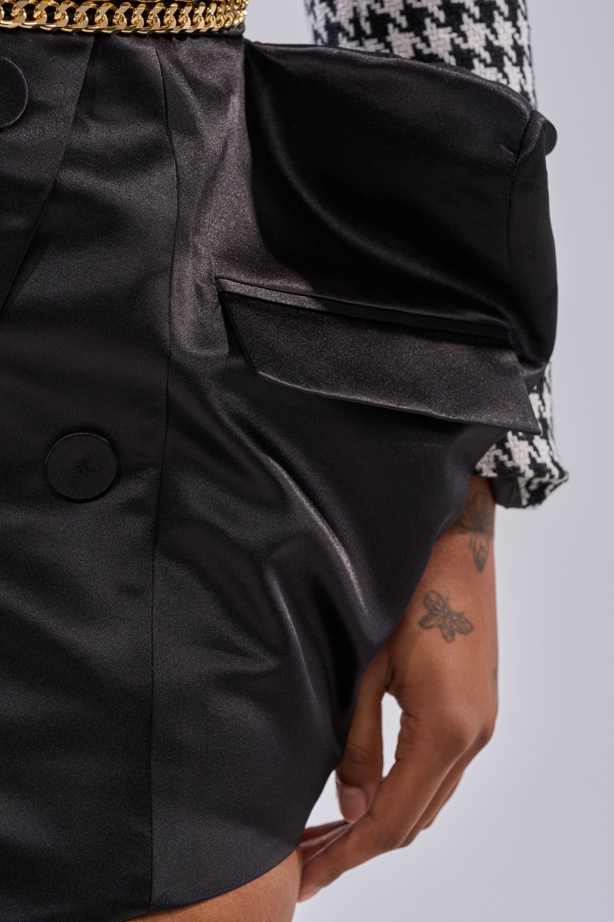 Close-up of a person wearing the ALL FOR IT MINI SKIRT, a black satin mini skirt with gold chain accents and a large pocket, paired with a houndstooth-patterned sleeve. Their butterfly tattooed hand rests beside their silhouette. 12