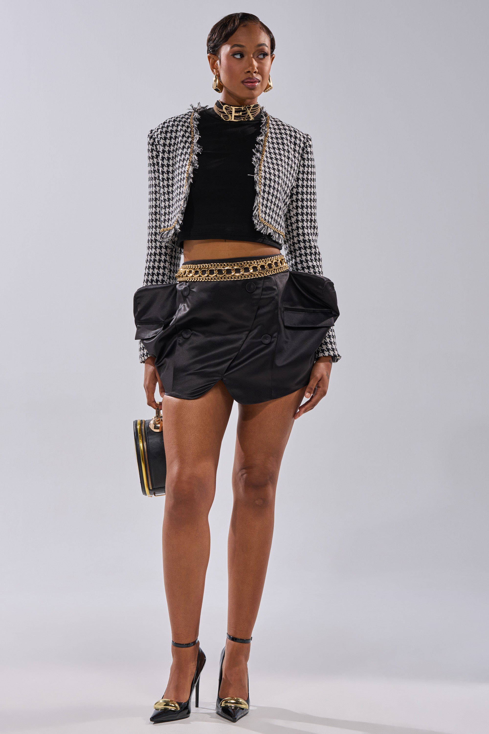 A woman stands against a plain background wearing the ALL FOR IT MINI SKIRT, a black crop top, a houndstooth jacket, black heels with gold chain accents, and holding a small black handbag. 11