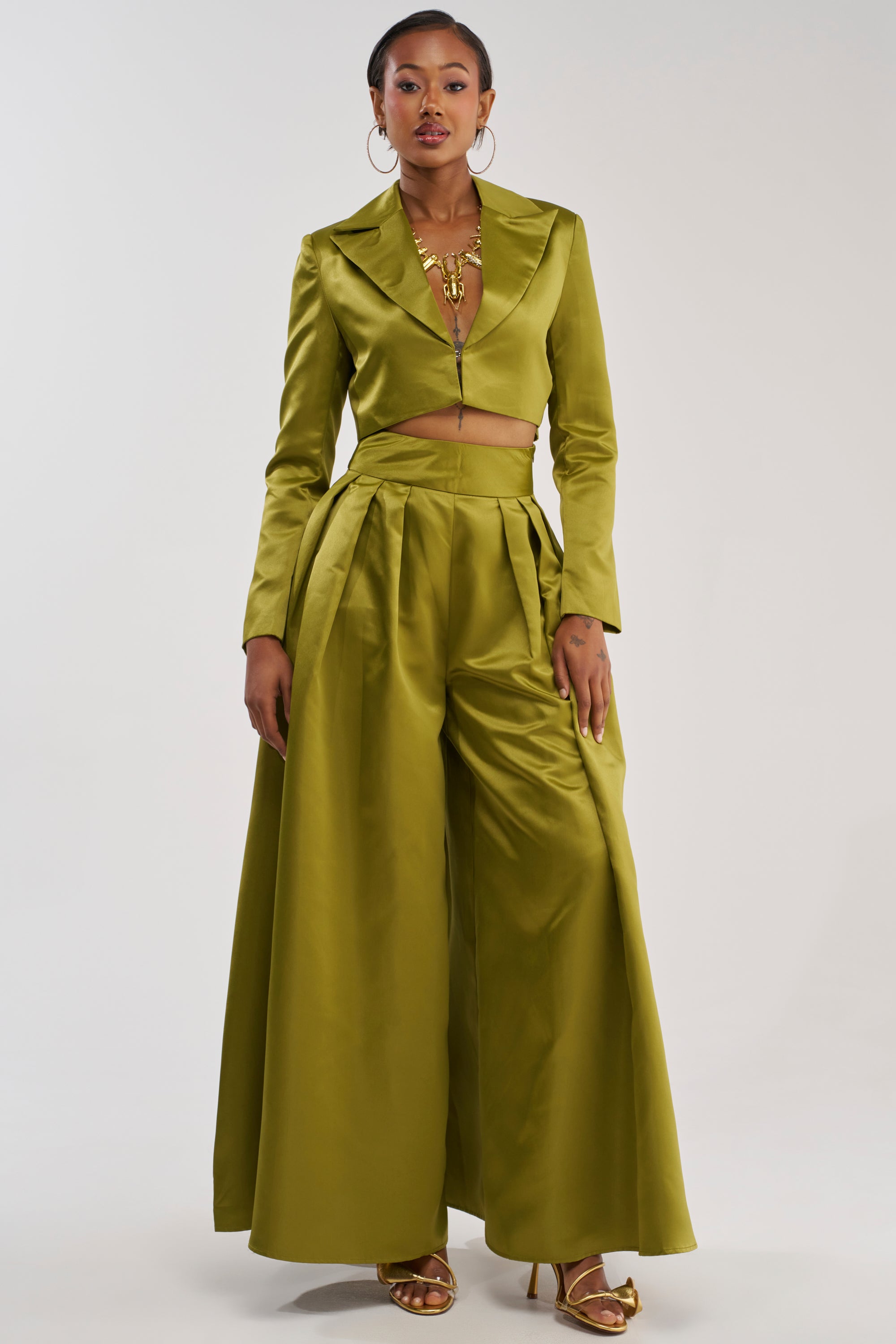 A woman models the LILA SATIN TROUSER IN CHARTREUSE, featuring a chic wide-leg silhouette in glossy satin. She pairs the statement trousers with a cropped blazer, gold heels, bold earrings, and layered necklaces as she stands confidently against a plain light background.