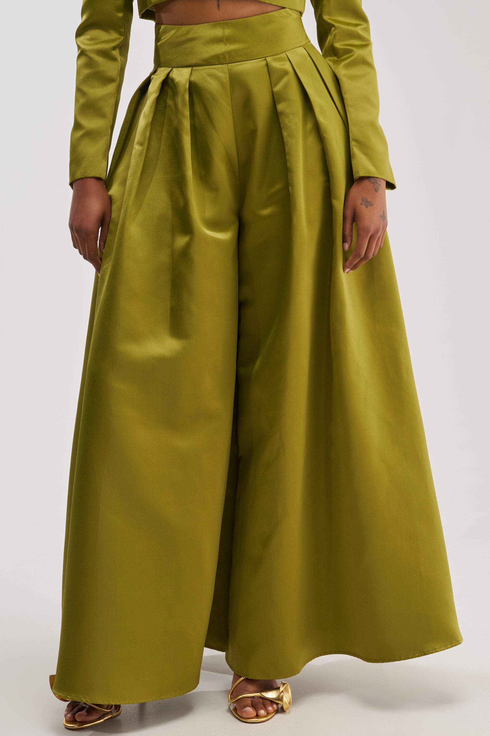 Wearing the LILA SATIN TROUSER IN CHARTREUSE, which features a high-waisted fit and bold wide-leg silhouette, a person pairs these striking satin pants with a matching long-sleeve top and gold open-toe heels, standing against a plain background.