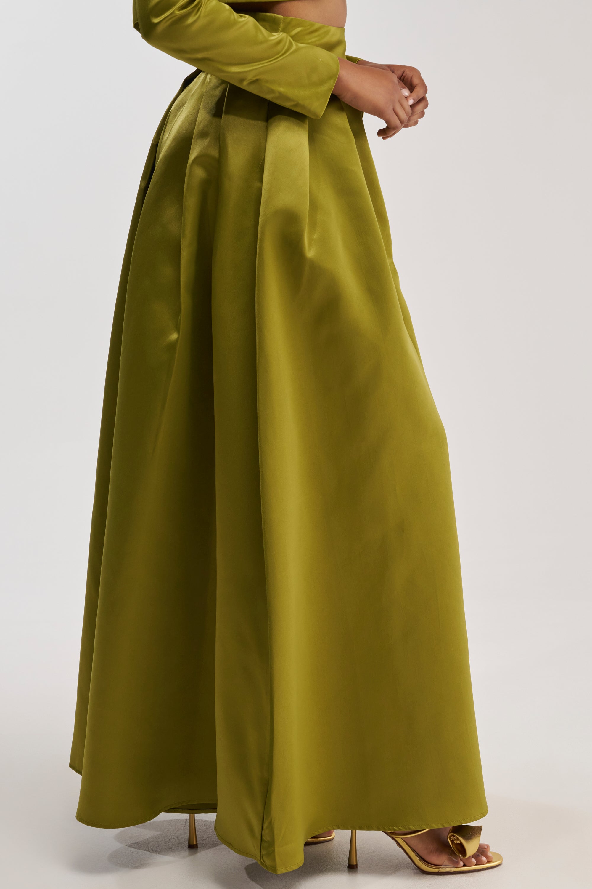 Dressed in the LILA SATIN TROUSER IN CHARTREUSE—featuring a long, high-waisted, wide-leg silhouette—a woman pairs the elegant bottoms with a matching crop top and strappy gold heels, standing against a plain light background for a chic formal evening look.