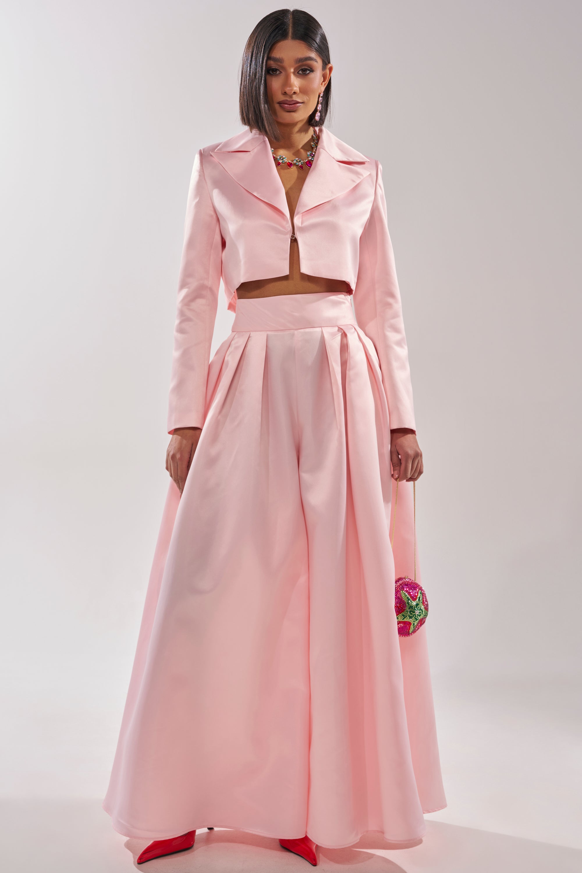 A woman wears the LILA SATIN WIDE LEG PANT with a silky blush pink cropped jacket, red pointed shoes, and holds a pink floral accessory. Her straight hair frames her confident expression. 0