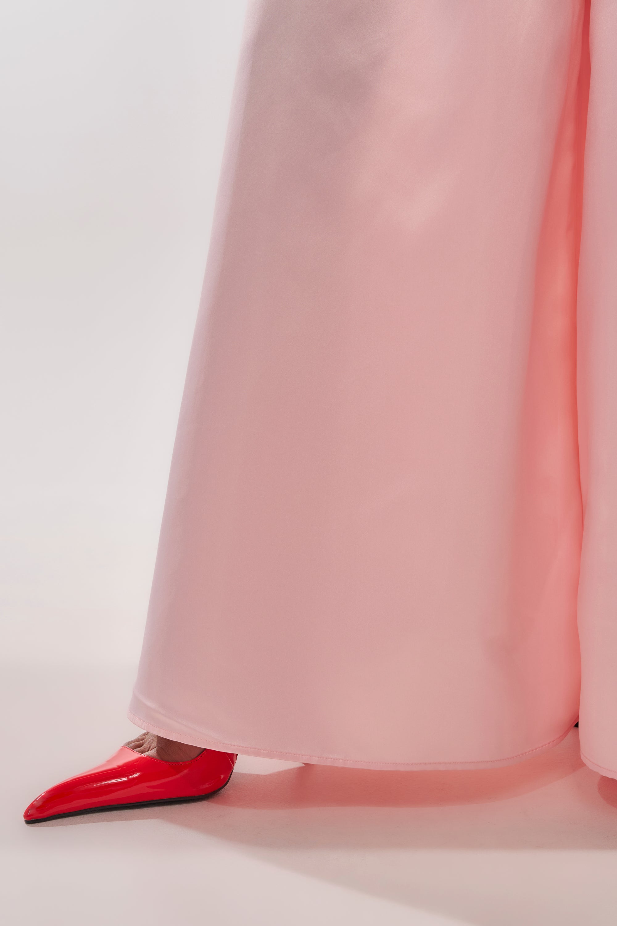 A person models the LILA SATIN WIDE LEG PANT in blush pink, standing with one foot forward to reveal a shiny red pointed-toe heel on a light background. 9