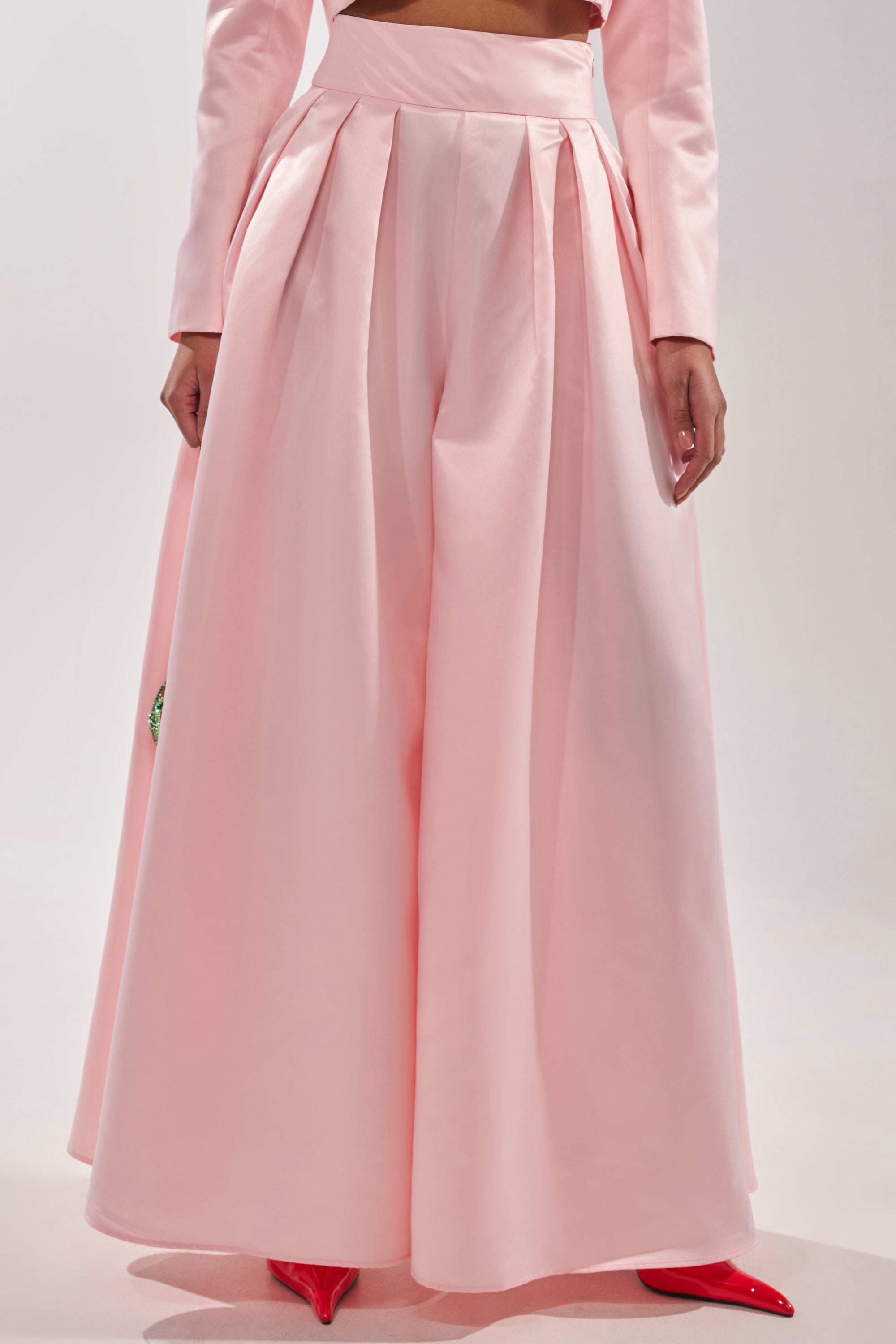 Someone stands against a plain light background wearing the LILA SATIN WIDE LEG PANT in blush pink with a high-rise waist and pleats, paired with matching pink suit sleeves and glossy red pointed-toe shoes. 1