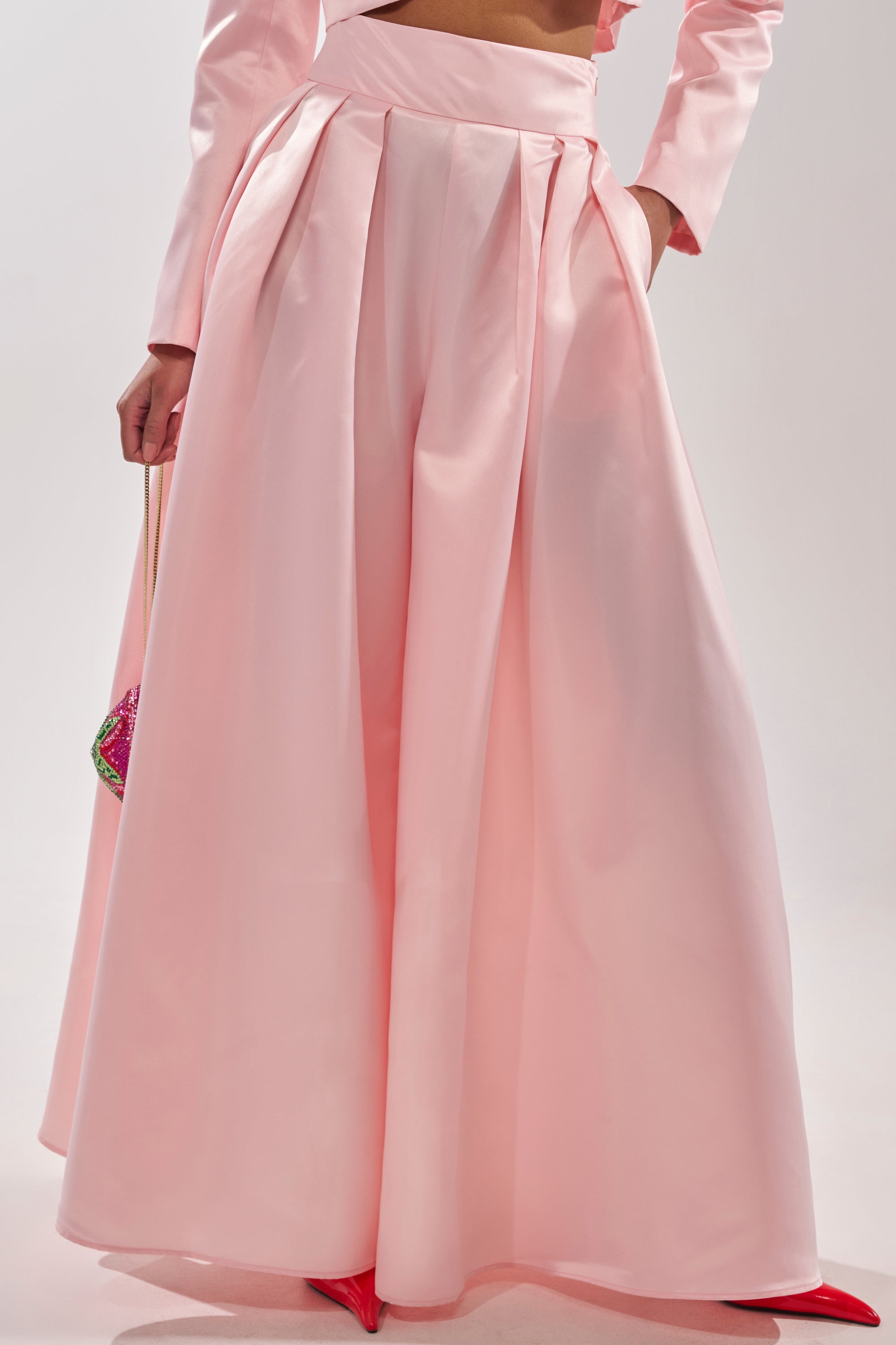 Wearing the LILA SATIN WIDE LEG PANT in blush pink with a high-rise waist and pleats, the lower body is shown holding a small purse and wearing bright red shoes. 2
