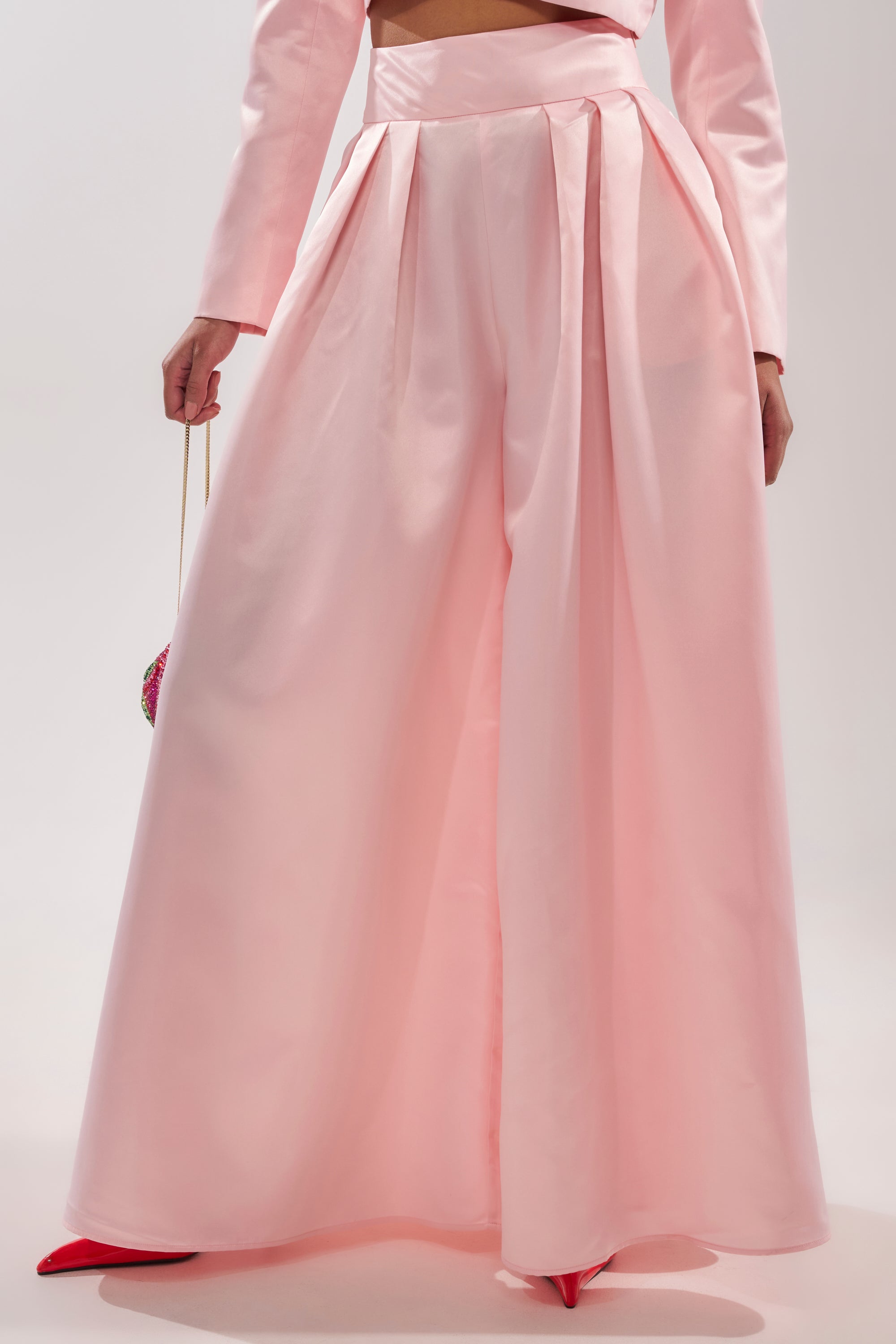 Wearing the LILA SATIN WIDE LEG PANT—high-rise, blush pink satin trousers with pleats and wide legs—paired with a matching long-sleeved top. Only the lower torso and legs are visible. 3