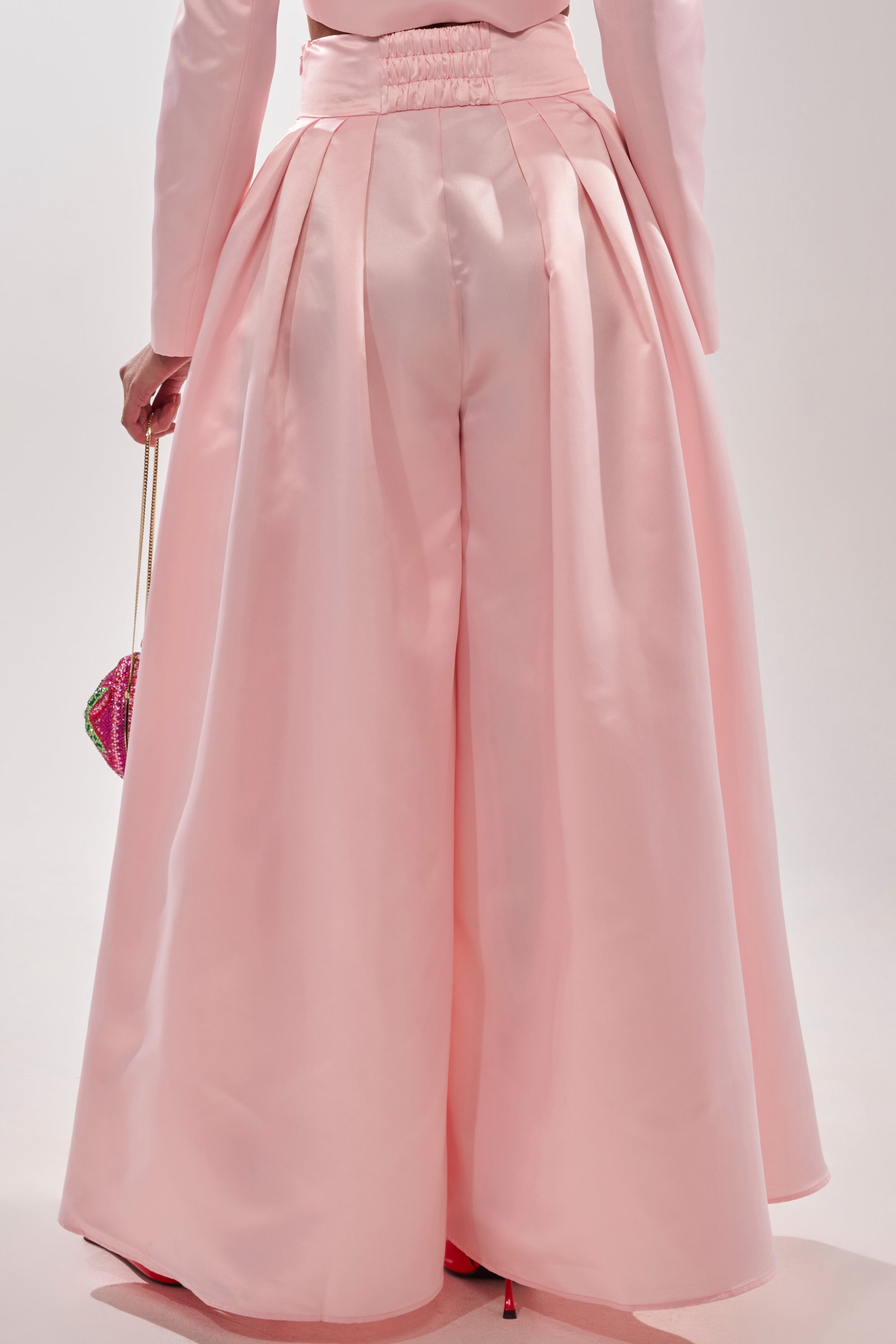 Someone photographed from behind wears the LILA SATIN WIDE LEG PANT in blush pink with pleats and holds a small, colorful beaded bag. 5