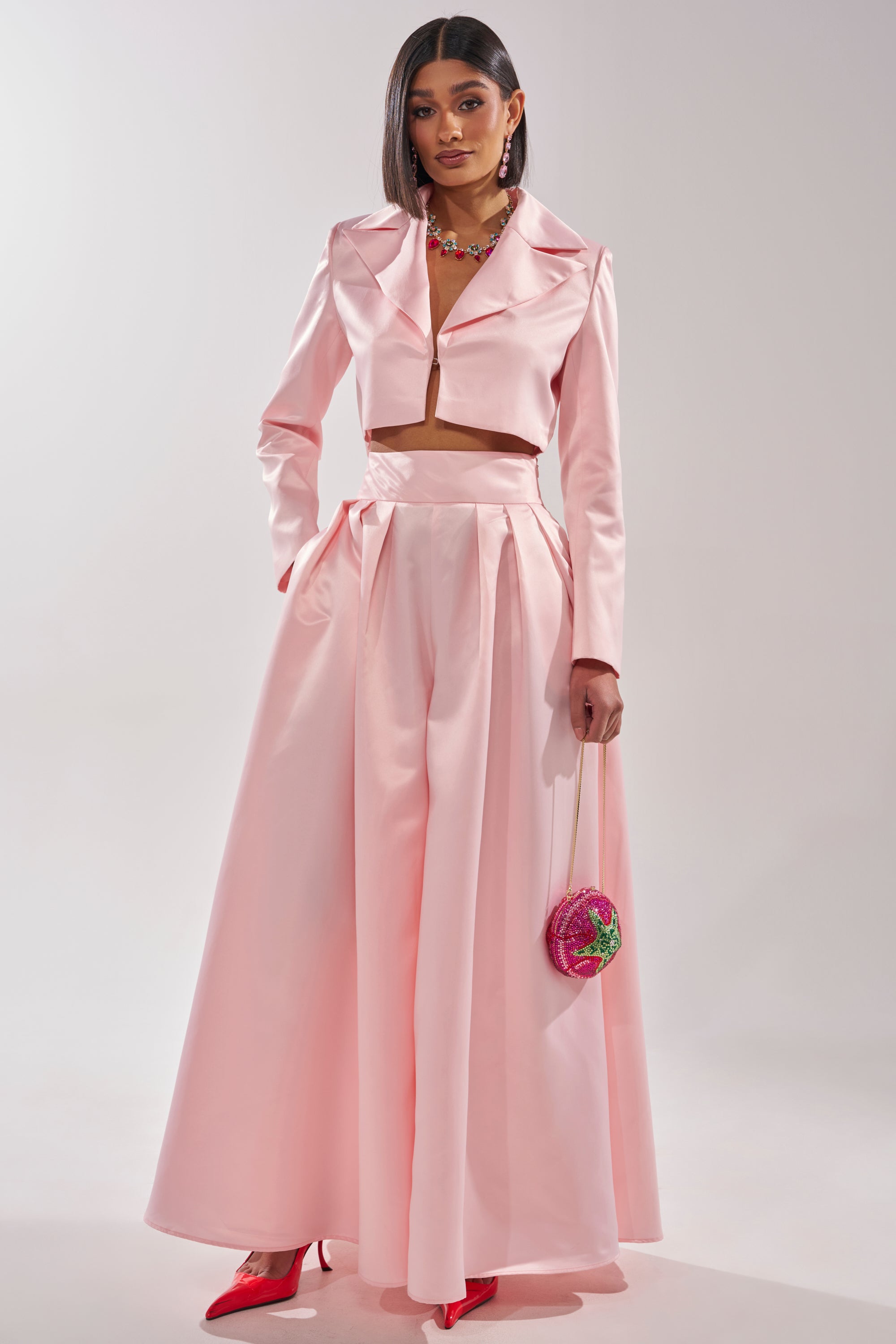 A woman wears the LILA SATIN WIDE LEG PANT in light pink with a high-rise waist, styled with a cropped blazer, red heels, long straight hair, and a small round pink purse. 6