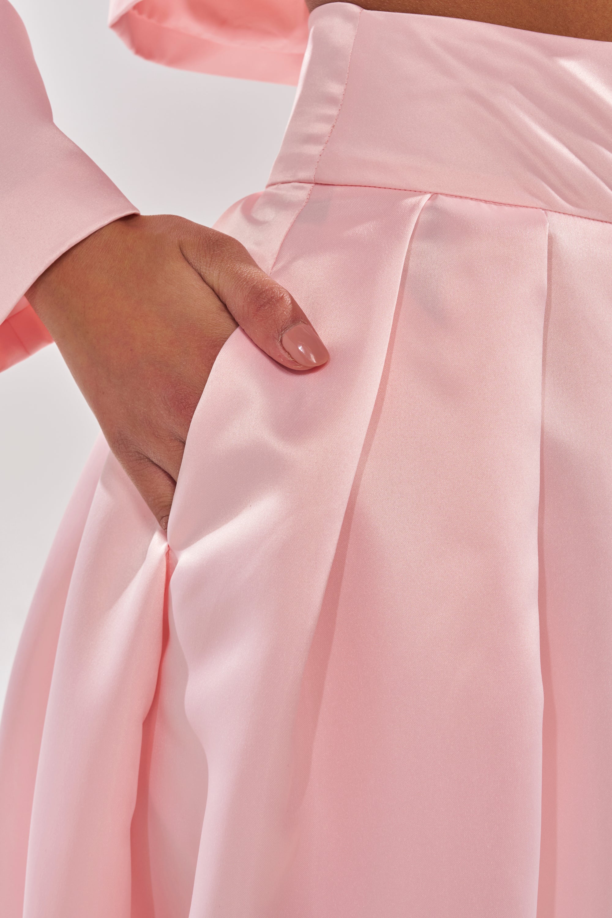 A person with neatly manicured nails slips their hand into the pocket of the blush pink LILA SATIN WIDE LEG PANT, featuring a high-rise waist and styled with a matching long-sleeve pink top. 8