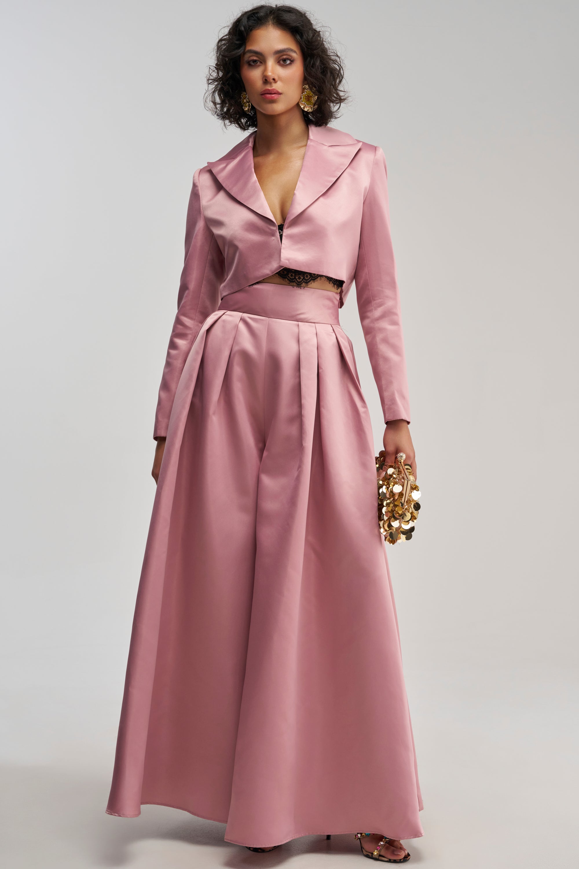 A woman stands wearing the LILA SATIN TROUSER IN MAUVE, styled as part of an elegant evening ensemble with a cropped jacket and a gold statement clutch.