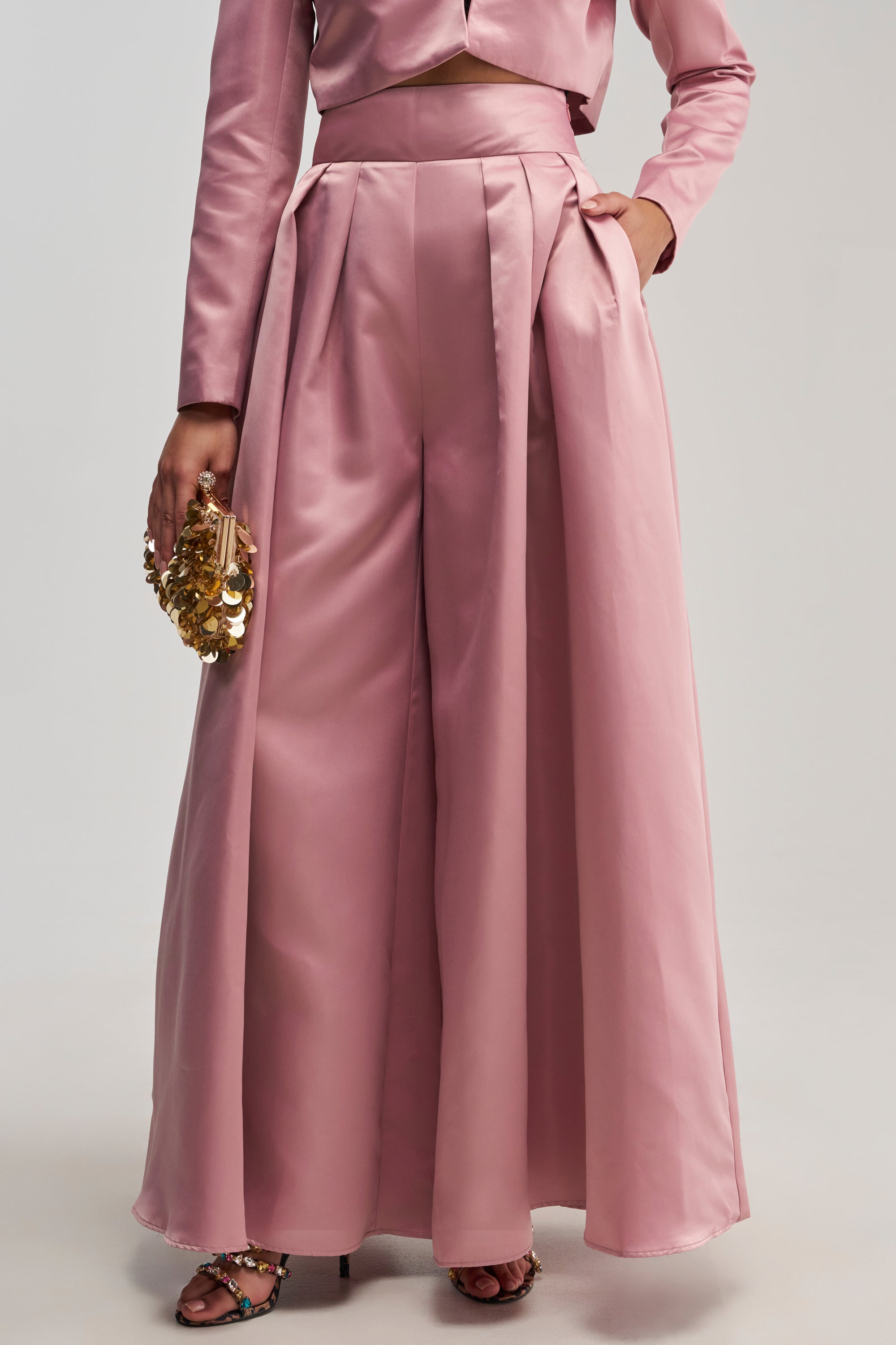 A person is shown from the waist down wearing the LILA SATIN TROUSER IN MAUVE—shiny pink wide-leg trousers—paired with a matching cropped blazer and holding a gold sequin clutch, standing against a plain background.