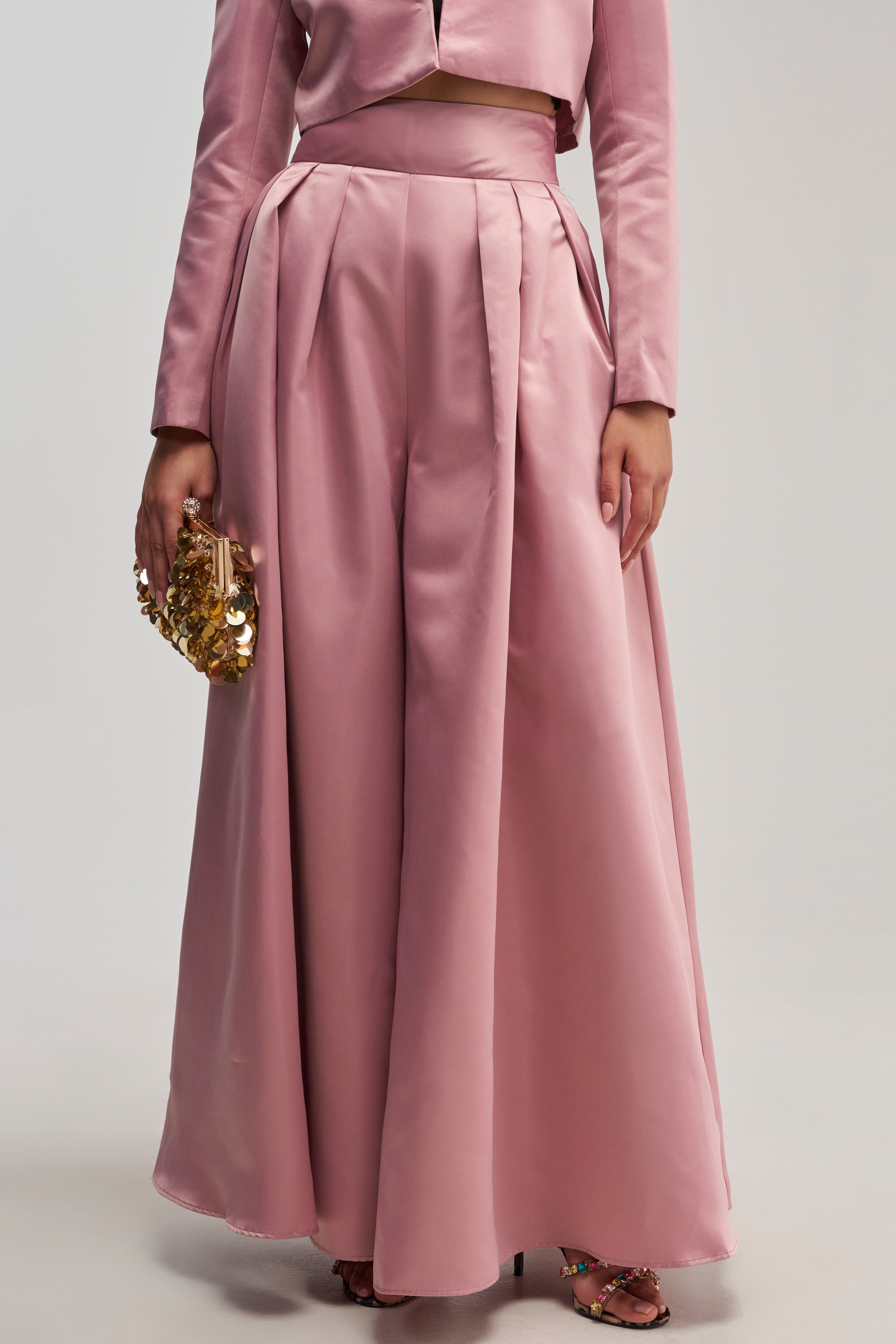 A person wearing the LILA SATIN TROUSER IN MAUVE, styled high-waisted with wide legs, pairs it with a matching cropped jacket, a gold beaded clutch, and open-toe heels, effortlessly showcasing chic evening wear against a plain background.