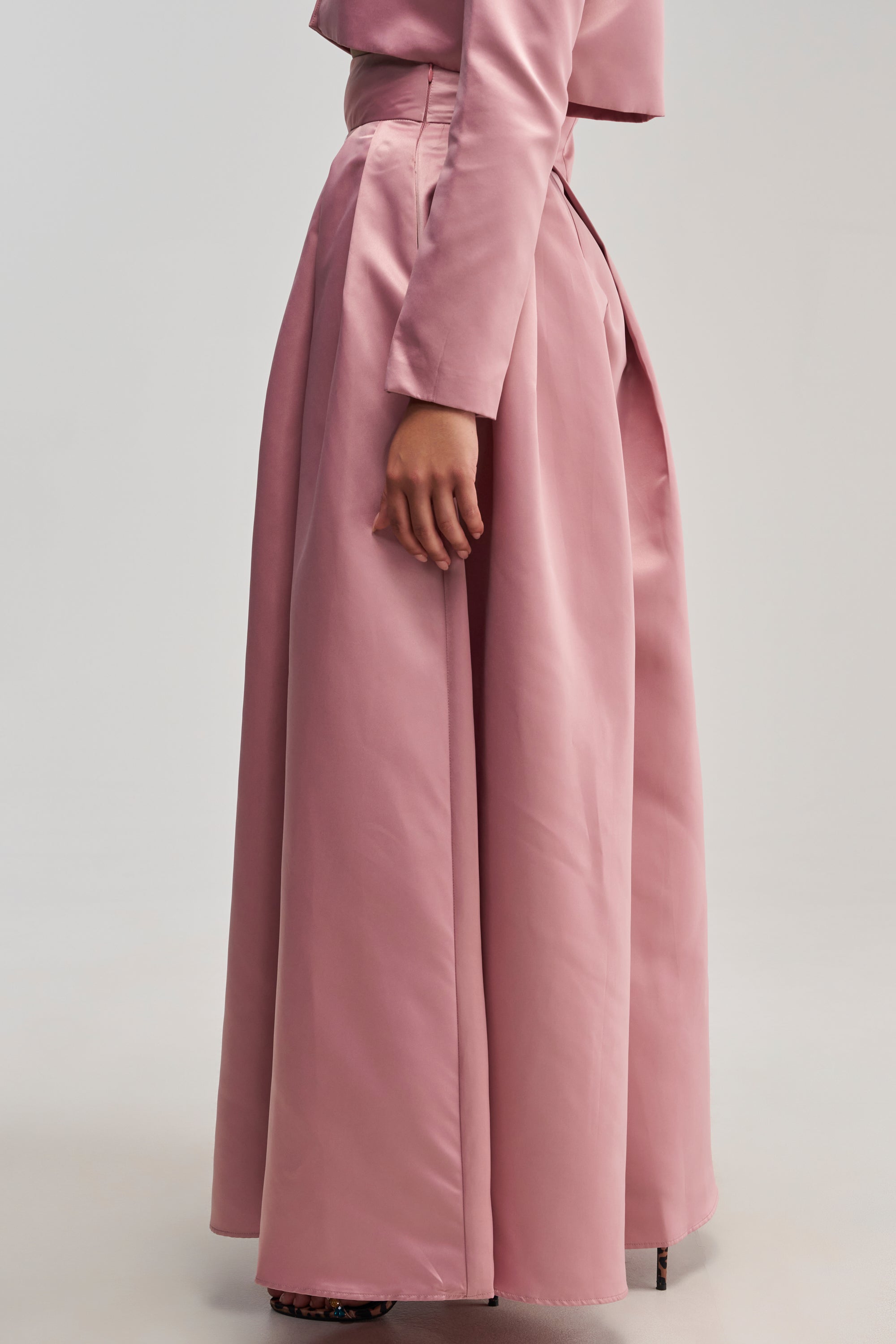 A person stands sideways, wearing the LILA SATIN TROUSER IN MAUVE, styled as elegant evening wear and paired with a matching pink top. Only their torso and legs are visible against a plain, light background.