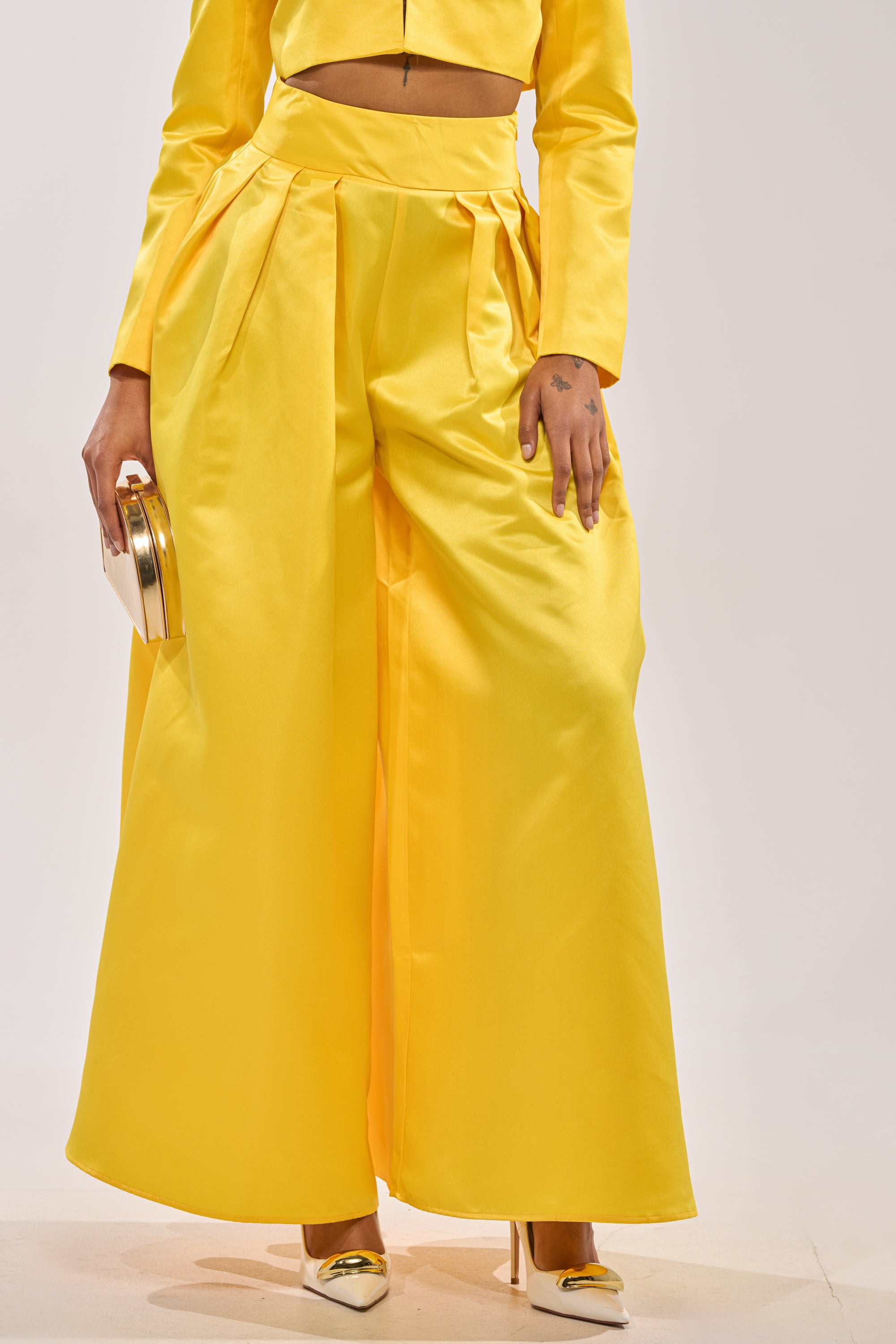 Wearing the BLOOMING WIDE LEG TROUSER in bright yellow with a matching long-sleeve top, gold clutch, and white heels with gold accents, the person stands against a light plain background. 1