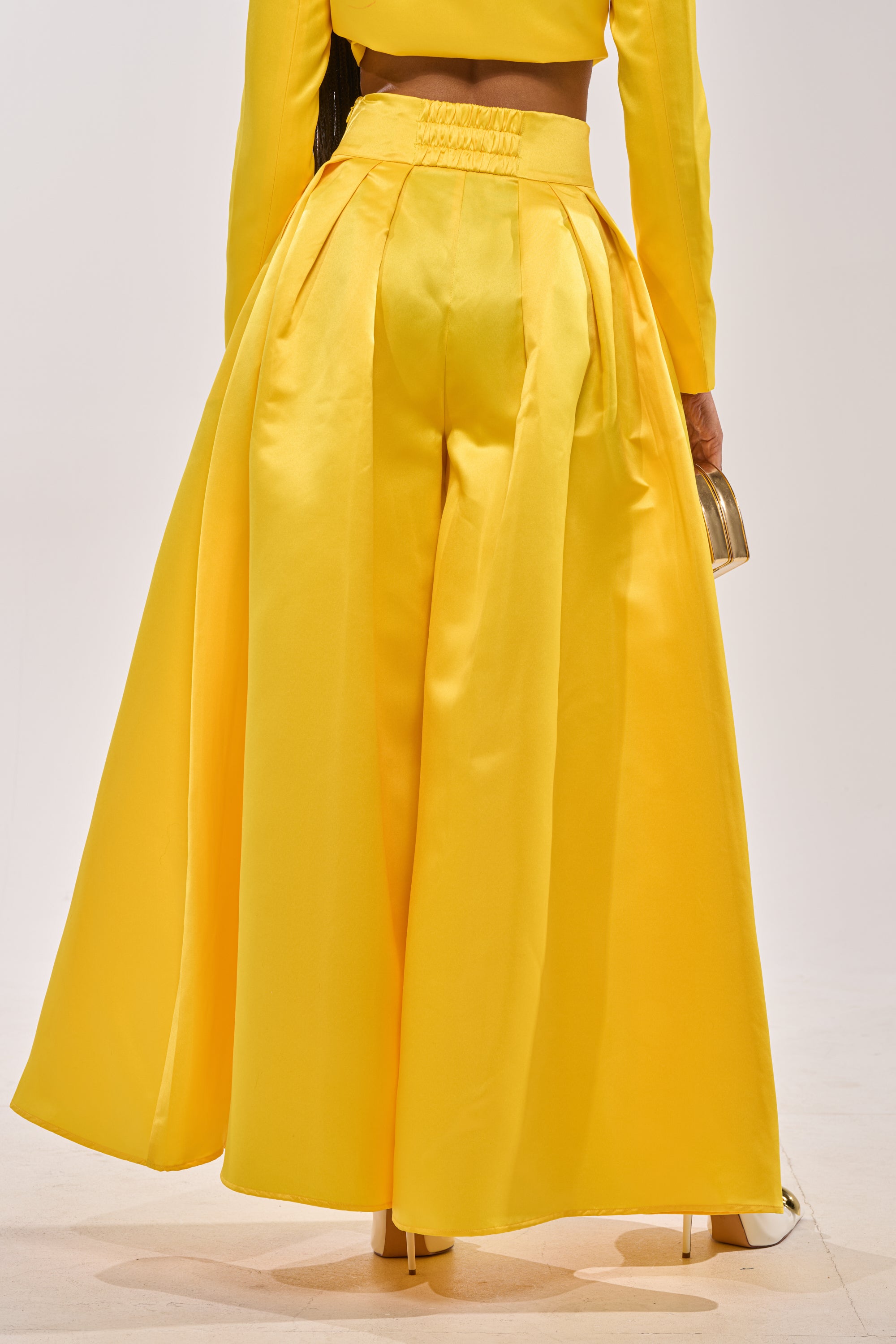 Someone stands with their back turned, showing only the lower half, in BLOOMING WIDE LEG TROUSER—bright yellow satin with a wide-leg cut—paired with a matching crop top and holding a small metallic handbag against a plain light background. 3