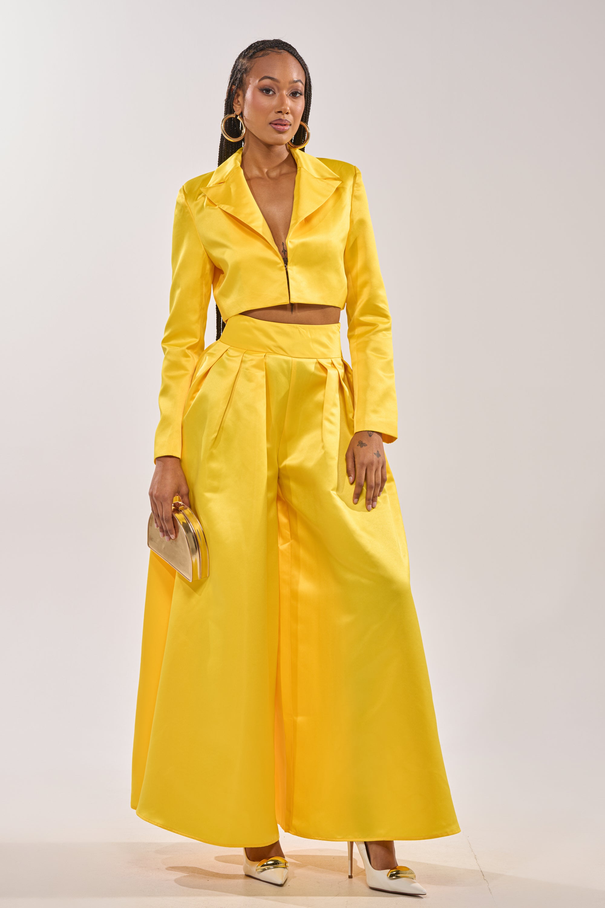 A woman stands confidently in the BLOOMING WIDE LEG TROUSER, a bright yellow high-waisted satin pant paired with a cropped jacket. She completes the look with a gold clutch, gold heels, and large hoop earrings against a plain background. 5