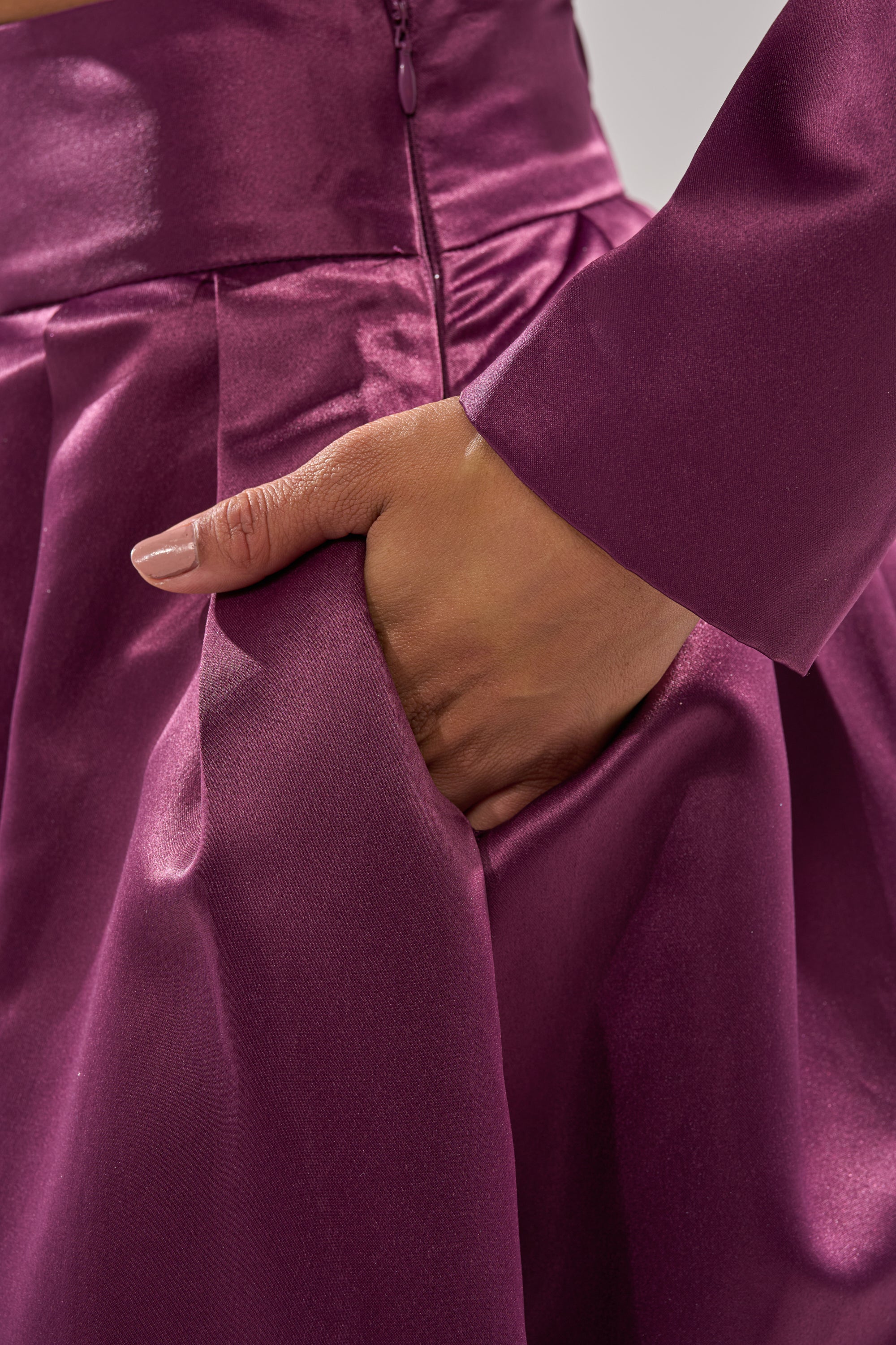 A close-up of a hand slipping into the pocket of the ON AND ON WIDE LEG TROUSER, crafted from shiny deep purple satin with a smooth texture and visible pleats.