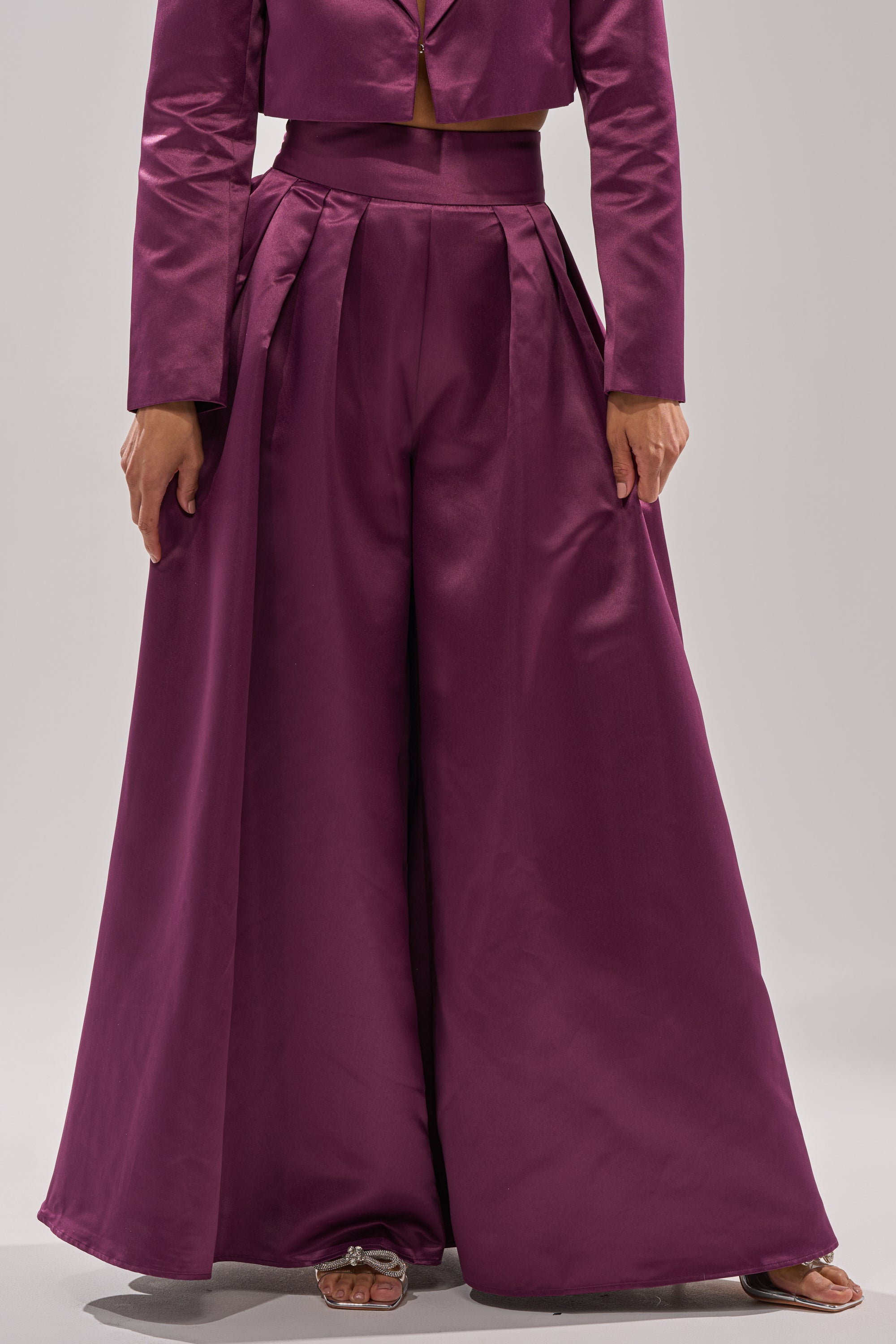 Wearing the ON AND ON WIDE LEG TROUSER in deep purple satin, a person stands against a neutral background. Only the lower half of their body is visible, highlighting the high-rise, wide-leg fit.