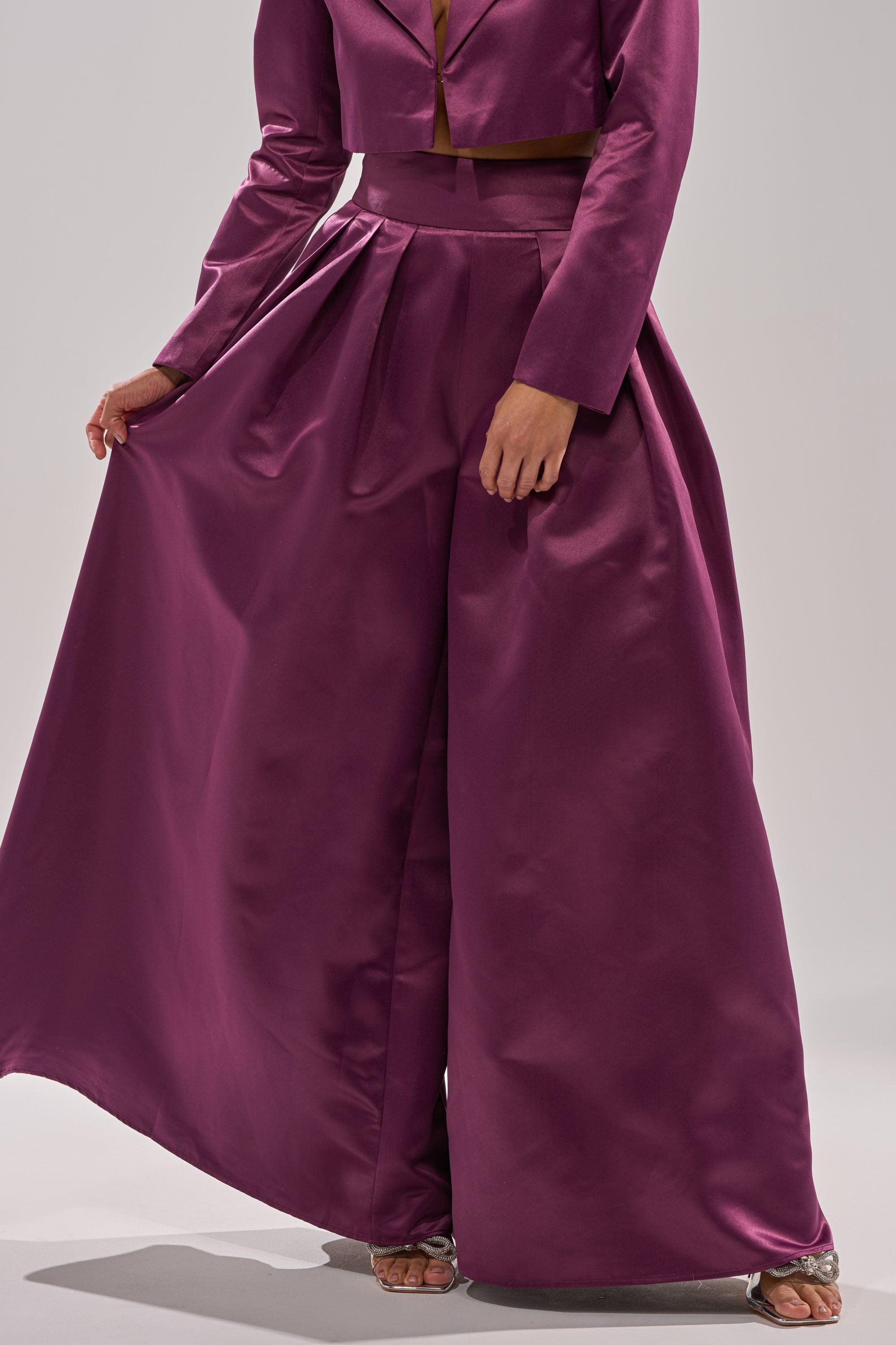 Wearing the ON AND ON WIDE LEG TROUSER, a person stands with one hand holding the flowing fabric. The look features a long-sleeved cropped magenta top and is finished with silver open-toe heels.