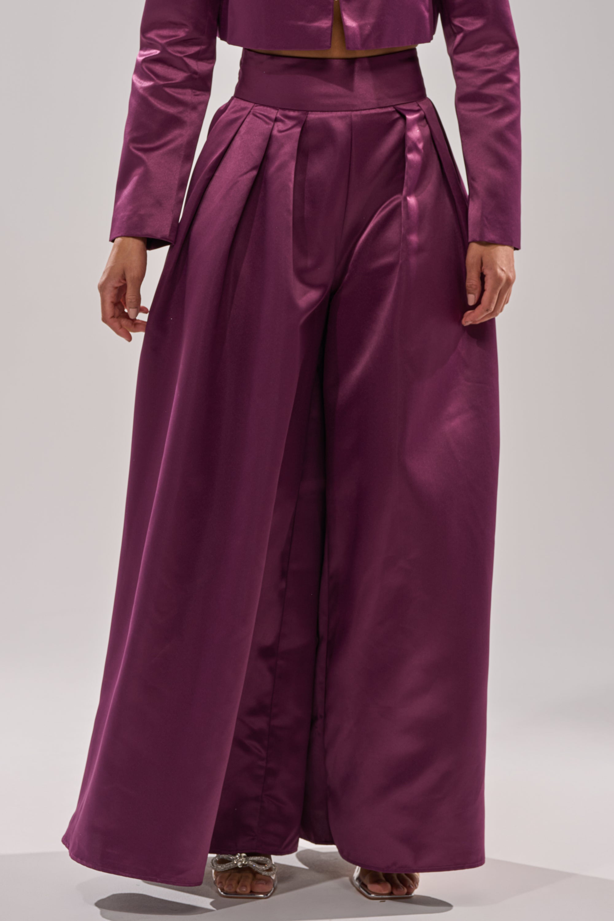 A person wearing the ON AND ON WIDE LEG TROUSER in magenta satin with a high-rise, wide-leg fit and pleats, paired with a matching long-sleeve crop top, shown from the waist down.