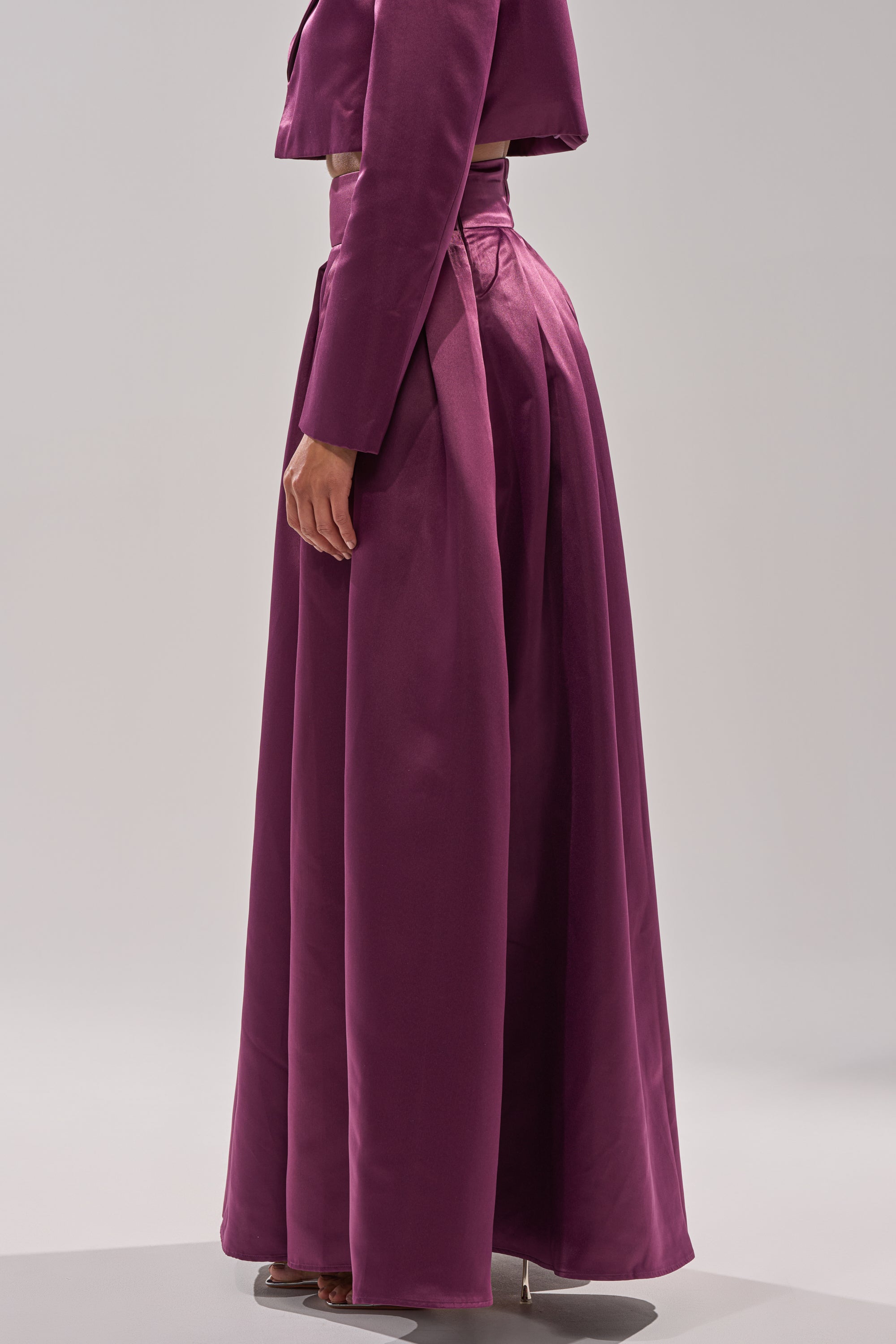 A person stands sideways against a plain light gray background, wearing the ON AND ON WIDE LEG TROUSER in deep magenta with pleats and a matching cropped jacket, styled high-waisted and floor-length.