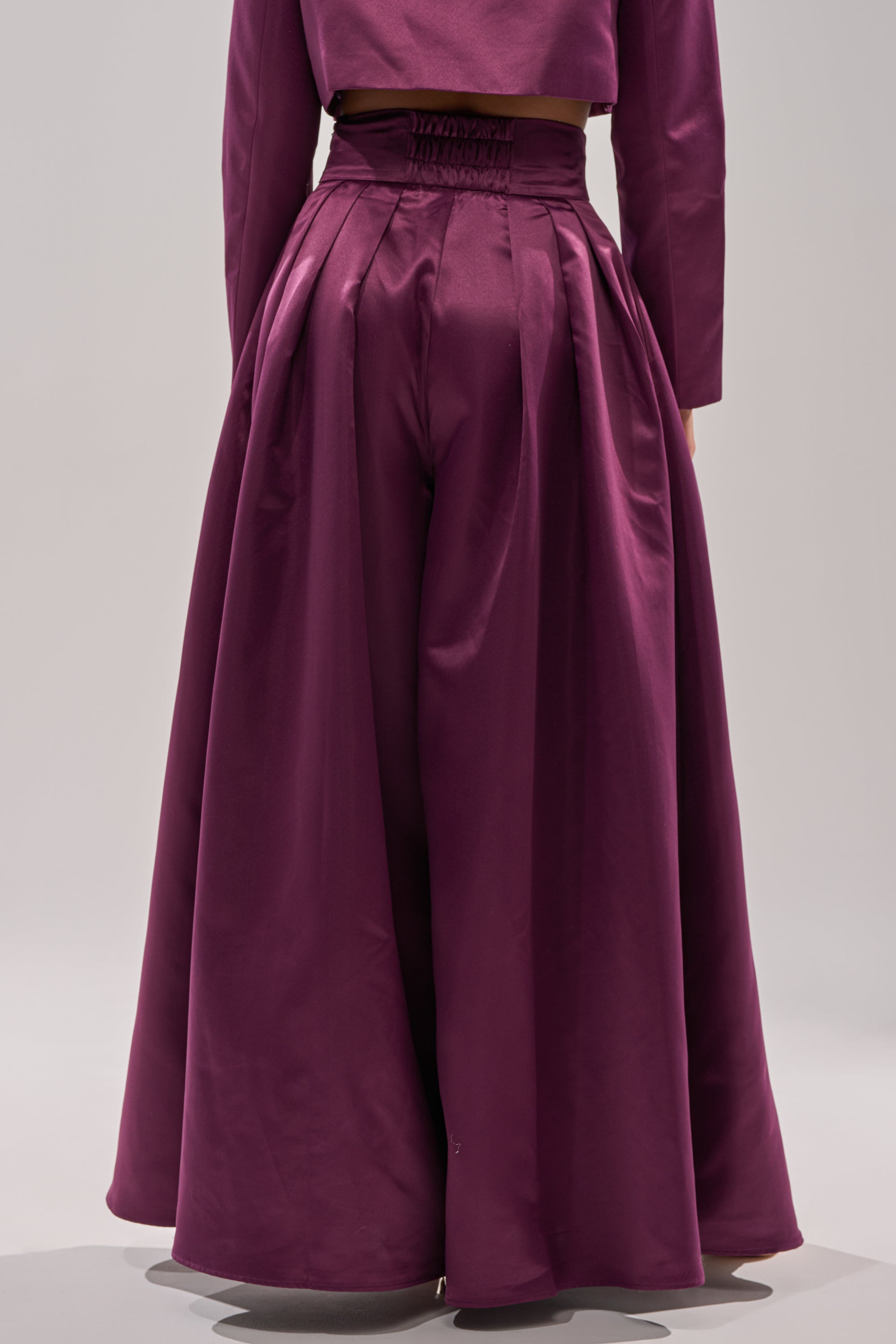 A person wears the ON AND ON WIDE LEG TROUSER, a high-rise, wide-leg satin pant in magenta, paired with a matching long-sleeve crop top, photographed from the waist down against a plain light background.
