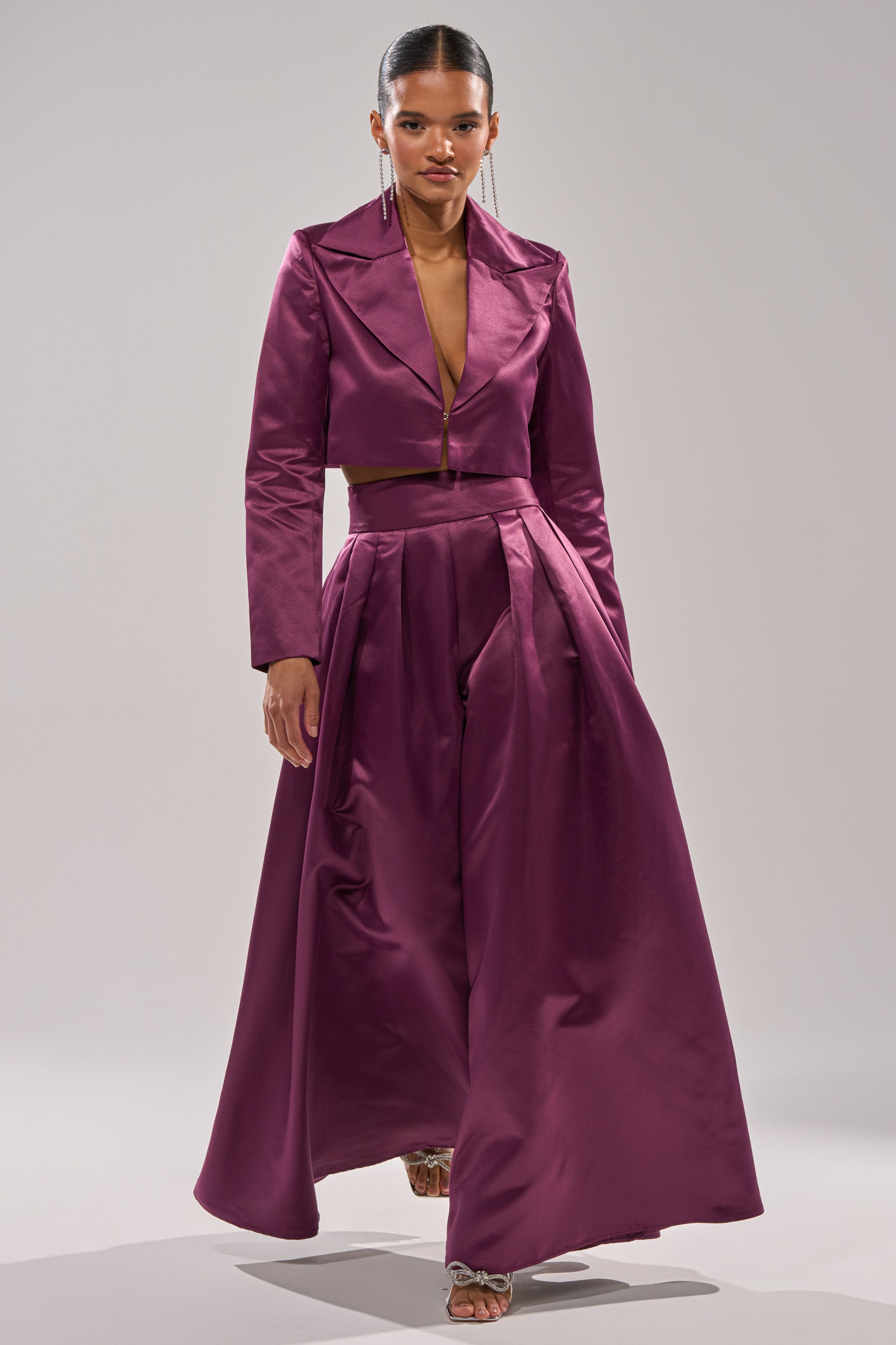 A woman strides confidently in the ON AND ON WIDE LEG TROUSER, part of a deep magenta satin set with a cropped V-neck jacket, styled with metallic heels and long earrings against a light backdrop.