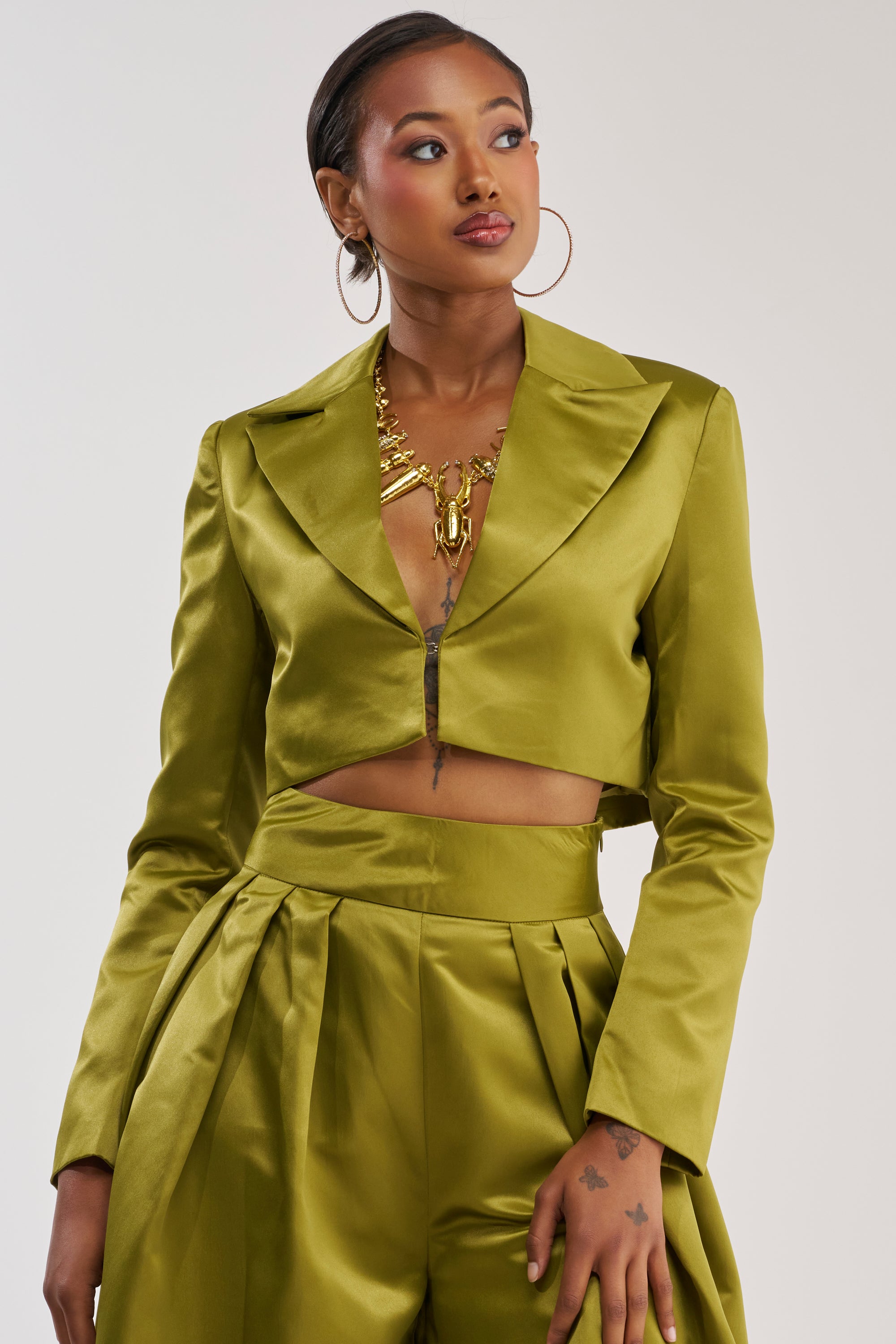 Wearing the LILA SATIN BLAZER IN CHARTREUSE, a woman embodies power dressing in this shiny olive-green satin blazer featuring a cropped fit and wide-legged pants, accessorized with gold jewelry and hoop earrings as she stands against a plain background.