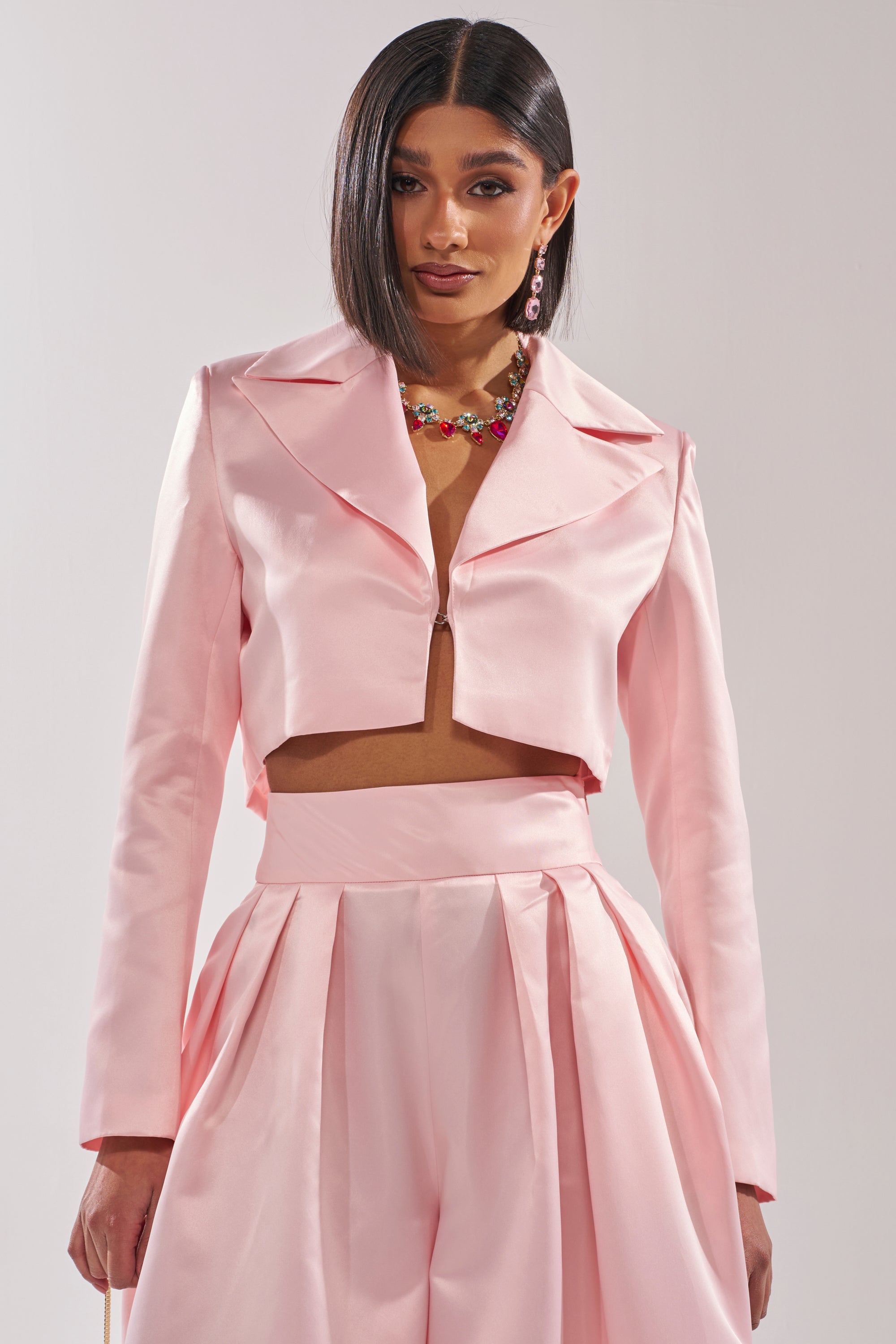 A person models the LILA SATIN BLAZER in blush pink, cropped and tailored, paired with matching high-waisted pleated pants, colorful necklace, and pink earrings against a simple light background. 0