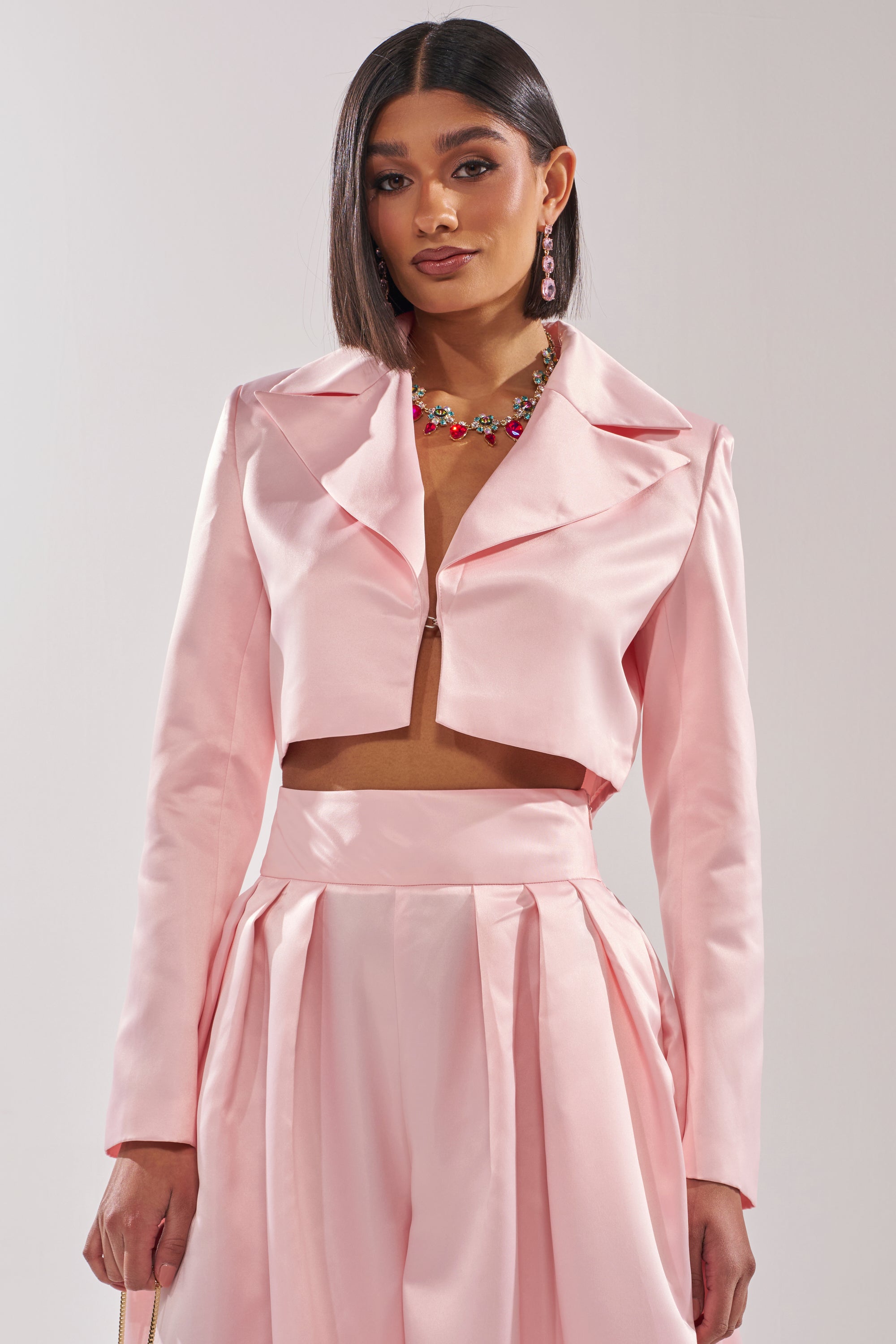 A woman with straight dark hair wears the LILA SATIN BLAZER in blush pink with matching high-waisted pleated pants, accessorized with a colorful necklace and pink earrings, standing against a plain light background. 1