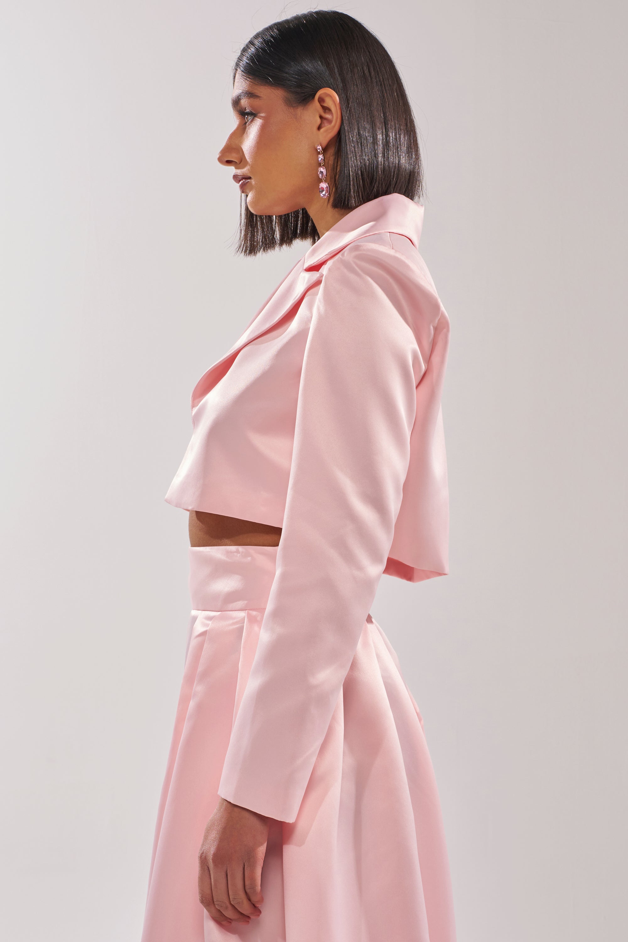 A woman with straight, shoulder-length dark hair wears the blush pink LILA SATIN BLAZER and matching high-waisted skirt, standing in profile against a plain light background. 2