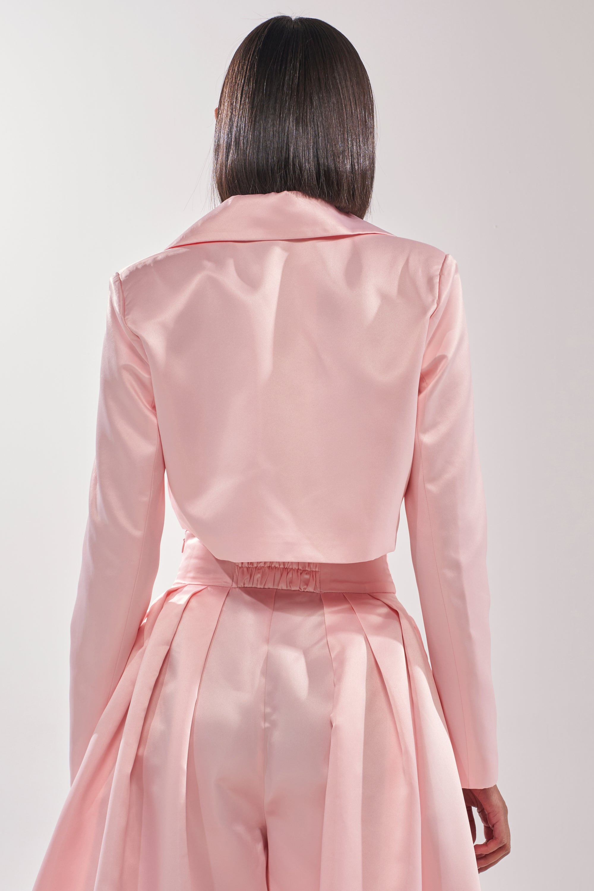 A person with straight, shoulder-length hair is seen from behind, wearing the LILA SATIN BLAZER in blush pink with high-waisted pleated trousers against a softly lit, plain background. 3