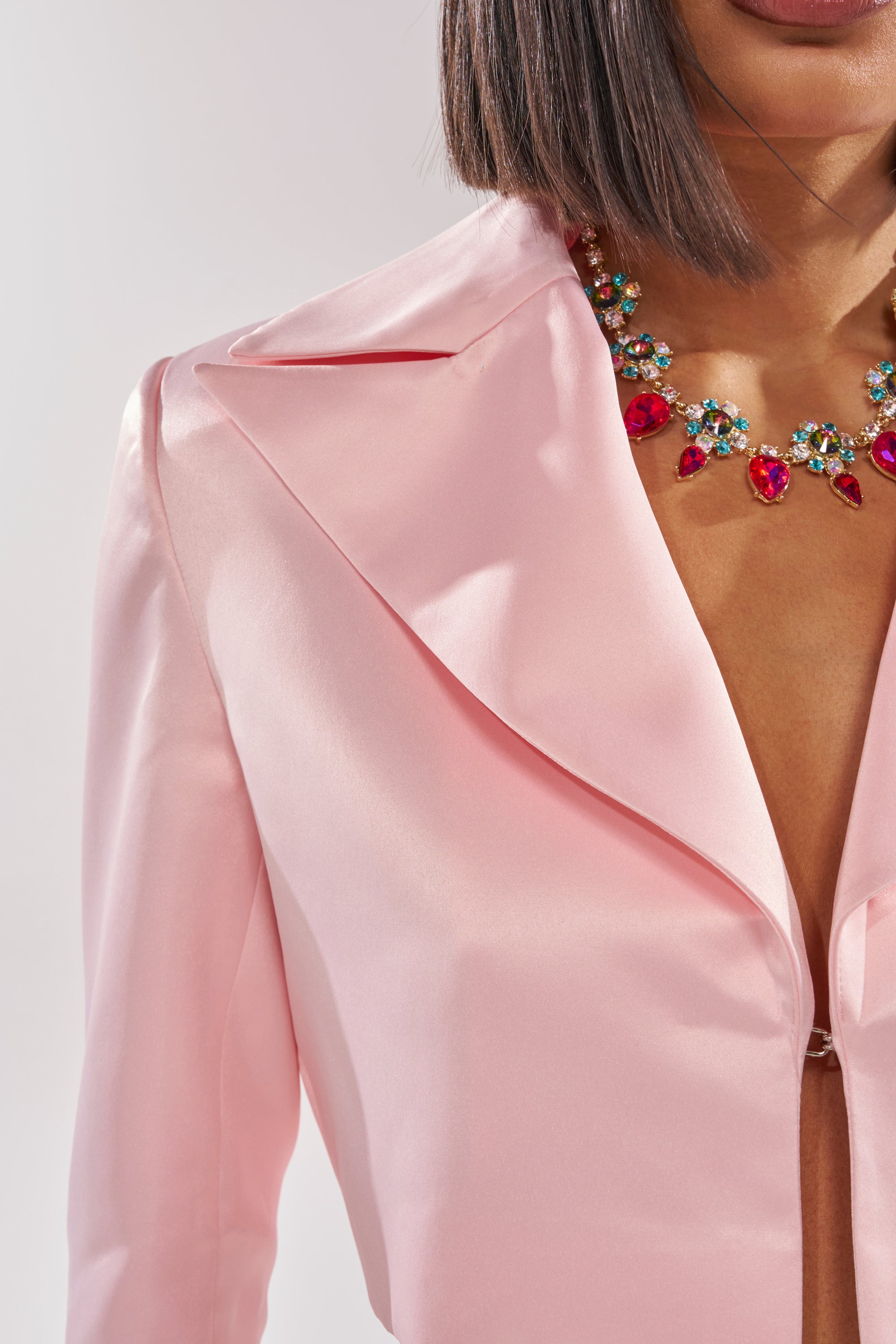 A woman models the LILA SATIN BLAZER in blush pink, featuring a cropped cut and deep neckline, paired with a vibrant gemstone necklace. Her straight, chin-length hair adds an edgy touch against the plain light background. 7
