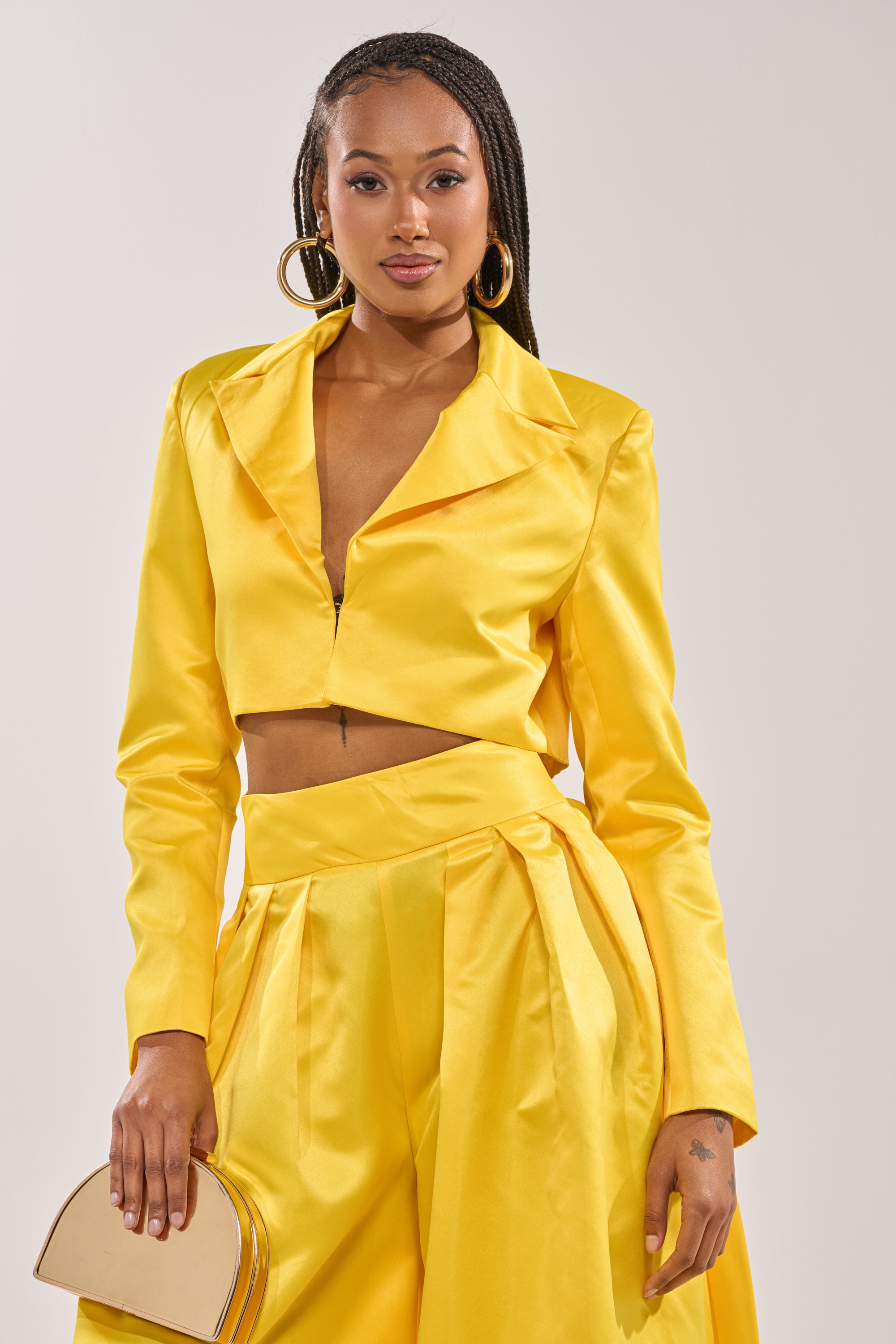 A woman wears the BLOOMING CROP SATIN BLAZER in yellow with matching high-waisted wide-leg pants, holding a small gold handbag. She has long braids, large hoop earrings, and a confident look. 0