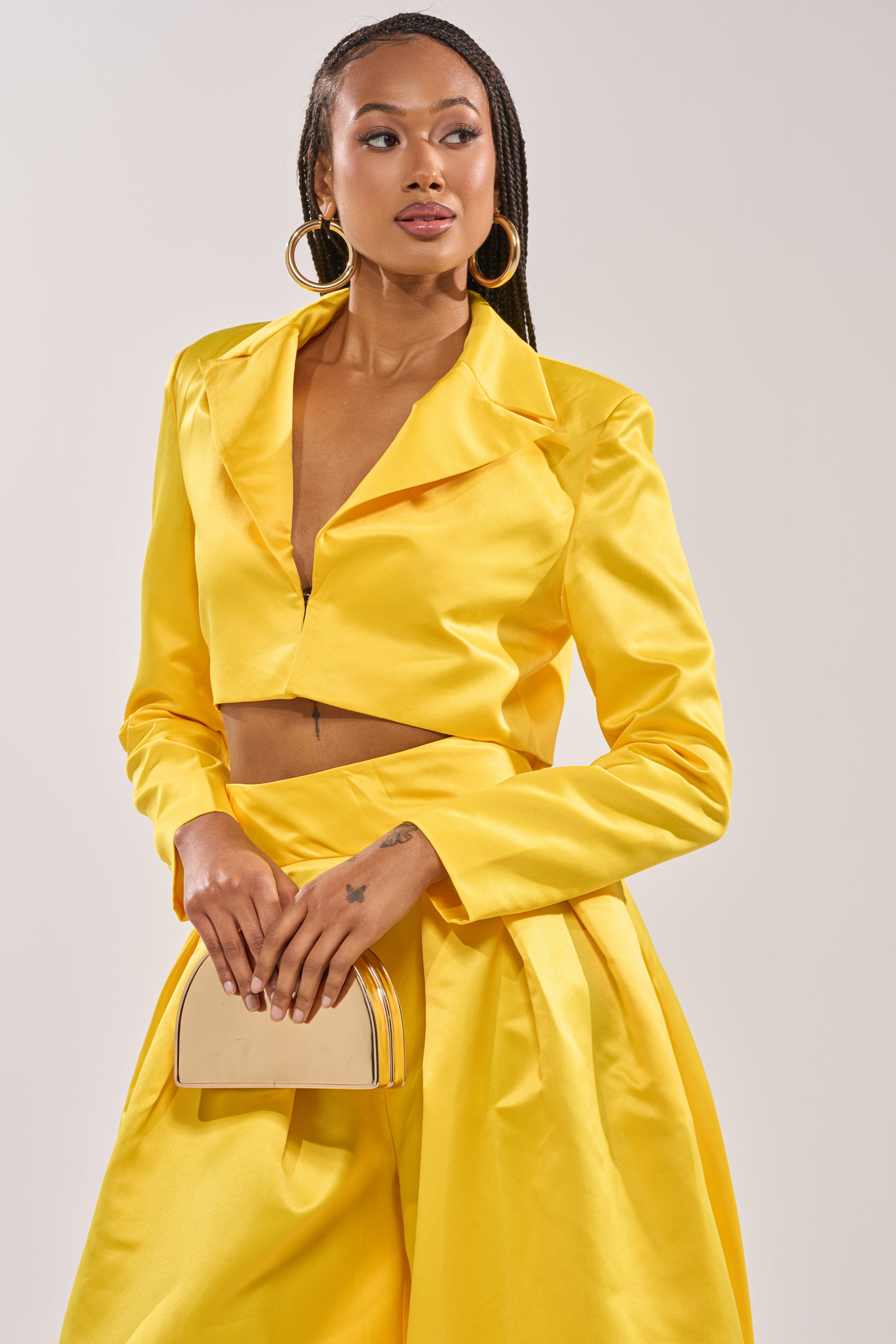 A woman exudes confidence in the BLOOMING CROP SATIN BLAZER and matching high-waisted skirt, accessorized with a cream clutch and hoop earrings—her braided hair completes this chic evening look. 1