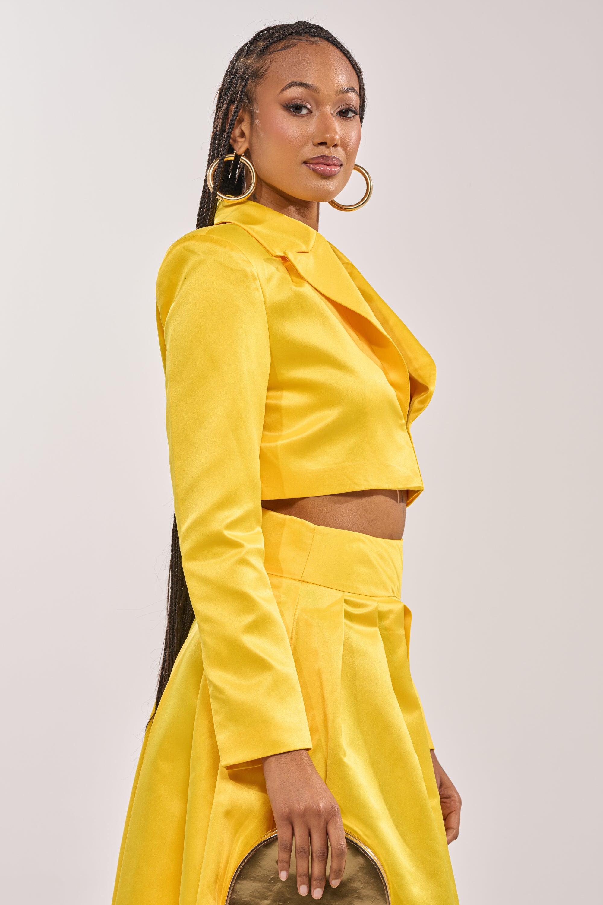 A woman with long braided hair poses confidently against a plain background, wearing the BLOOMING CROP SATIN BLAZER and a matching high-waisted skirt while holding a gold clutch. 2