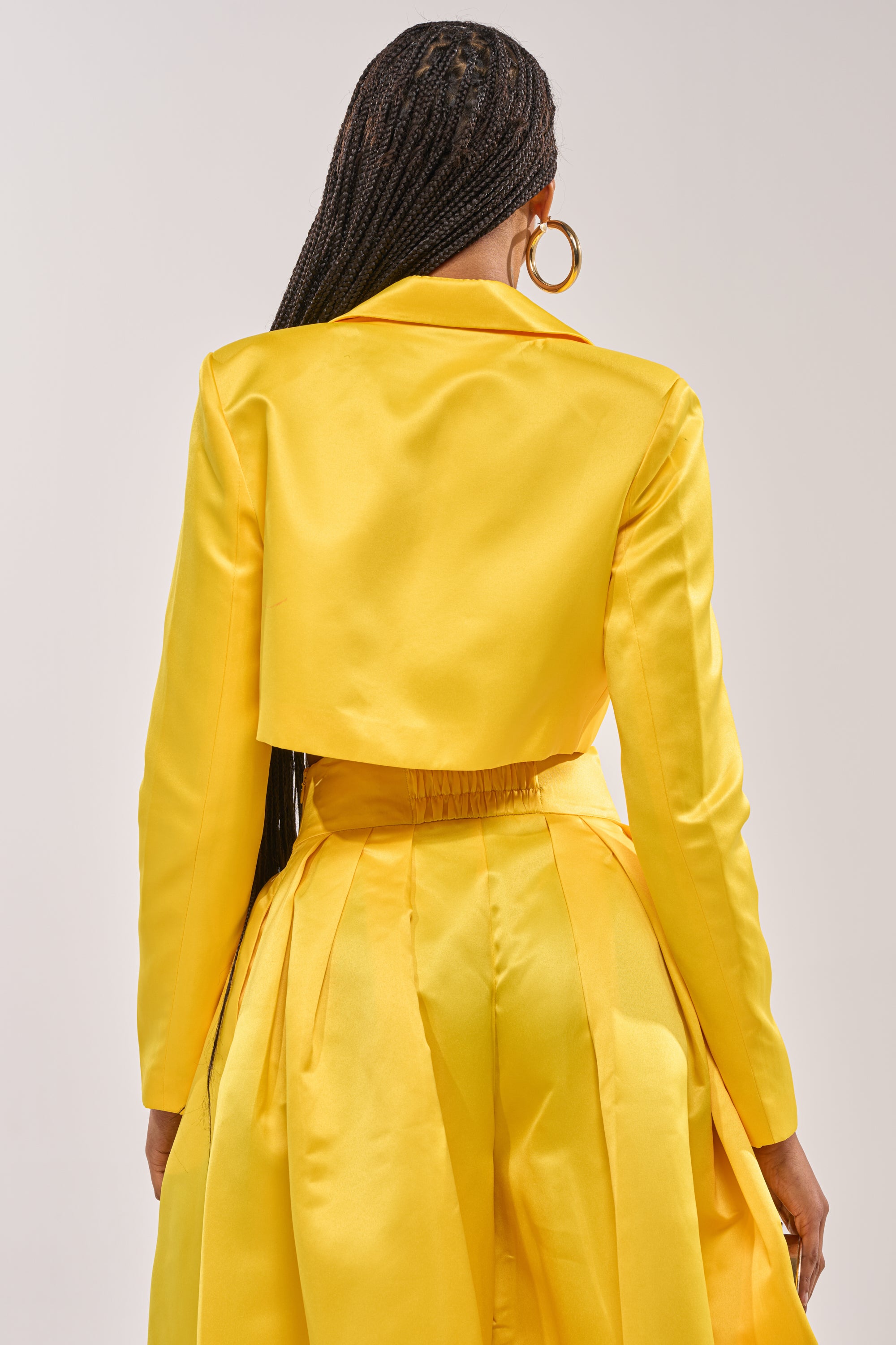 A woman with long braids and hoop earrings is shown from the back against a neutral background, wearing the BLOOMING CROP SATIN BLAZER paired with a matching pleated skirt. 3