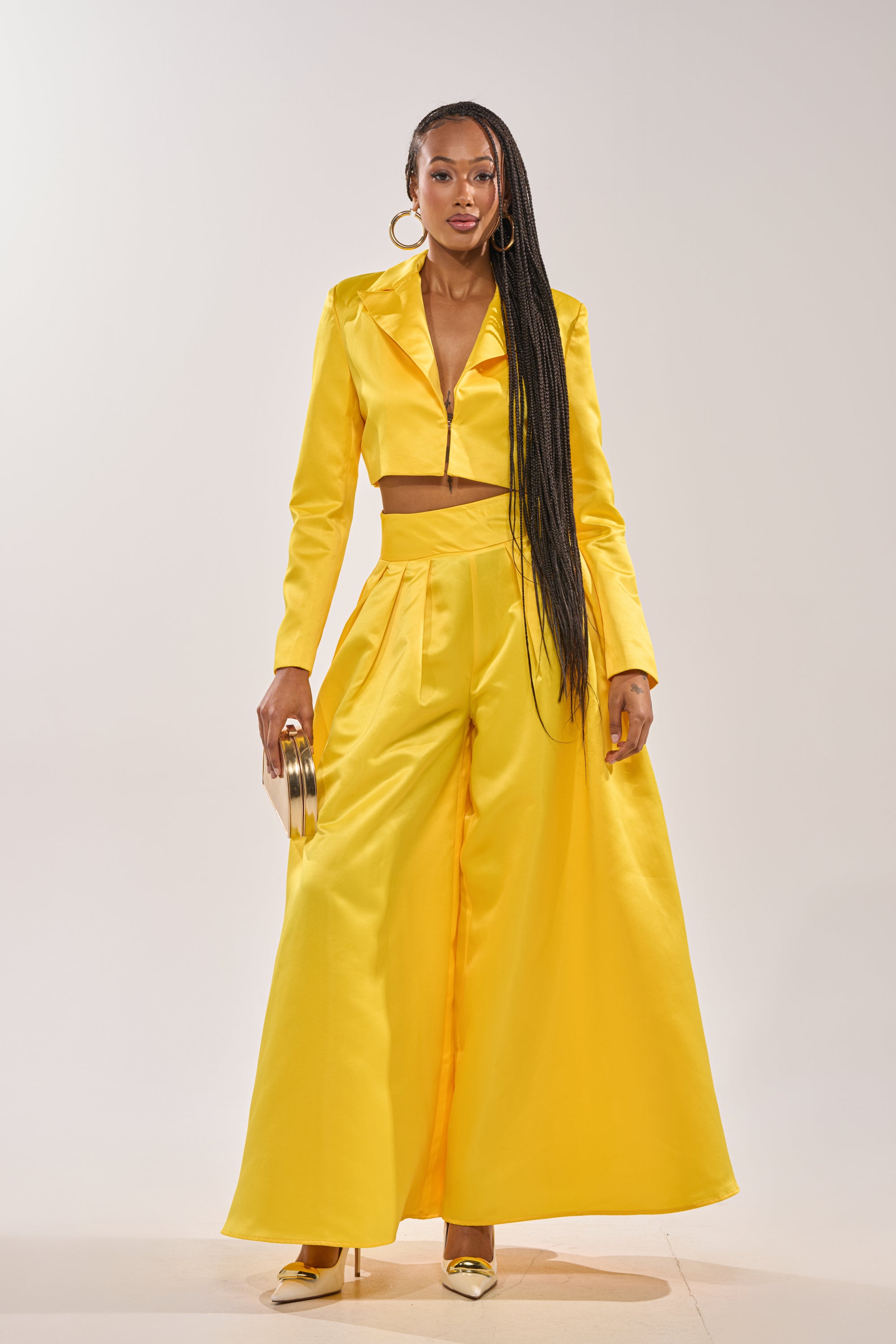 A woman stands confidently in the BLOOMING CROP SATIN BLAZER and matching wide-leg pants, holding a round gold clutch, wearing yellow heels, with long braided hair and hoop earrings. 4