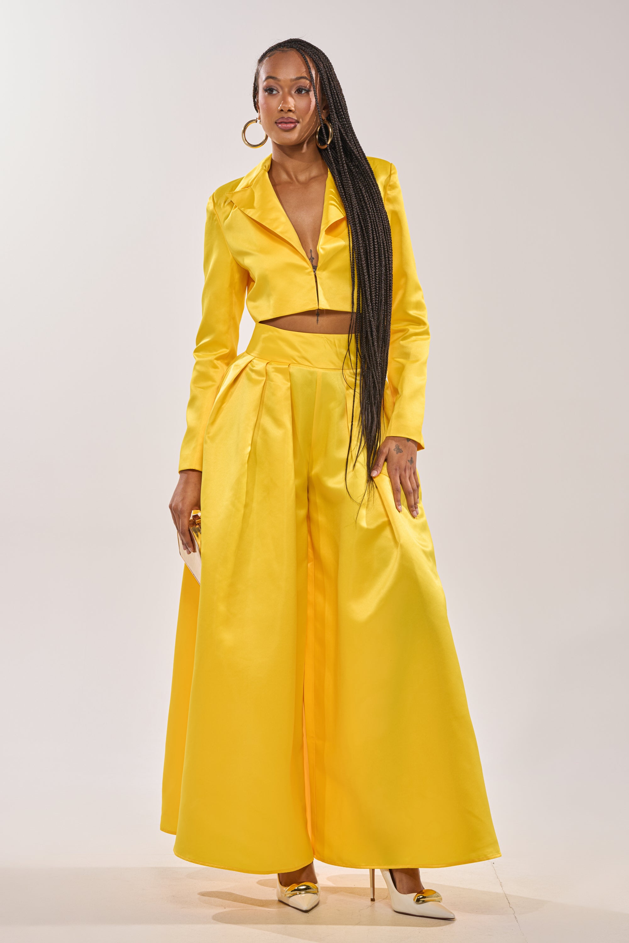 A woman stands confidently in the BLOOMING CROP SATIN BLAZER with matching wide-leg high-waisted pants and yellow heels, her long braided hair flowing as she poses with a small clutch against a plain background. 5