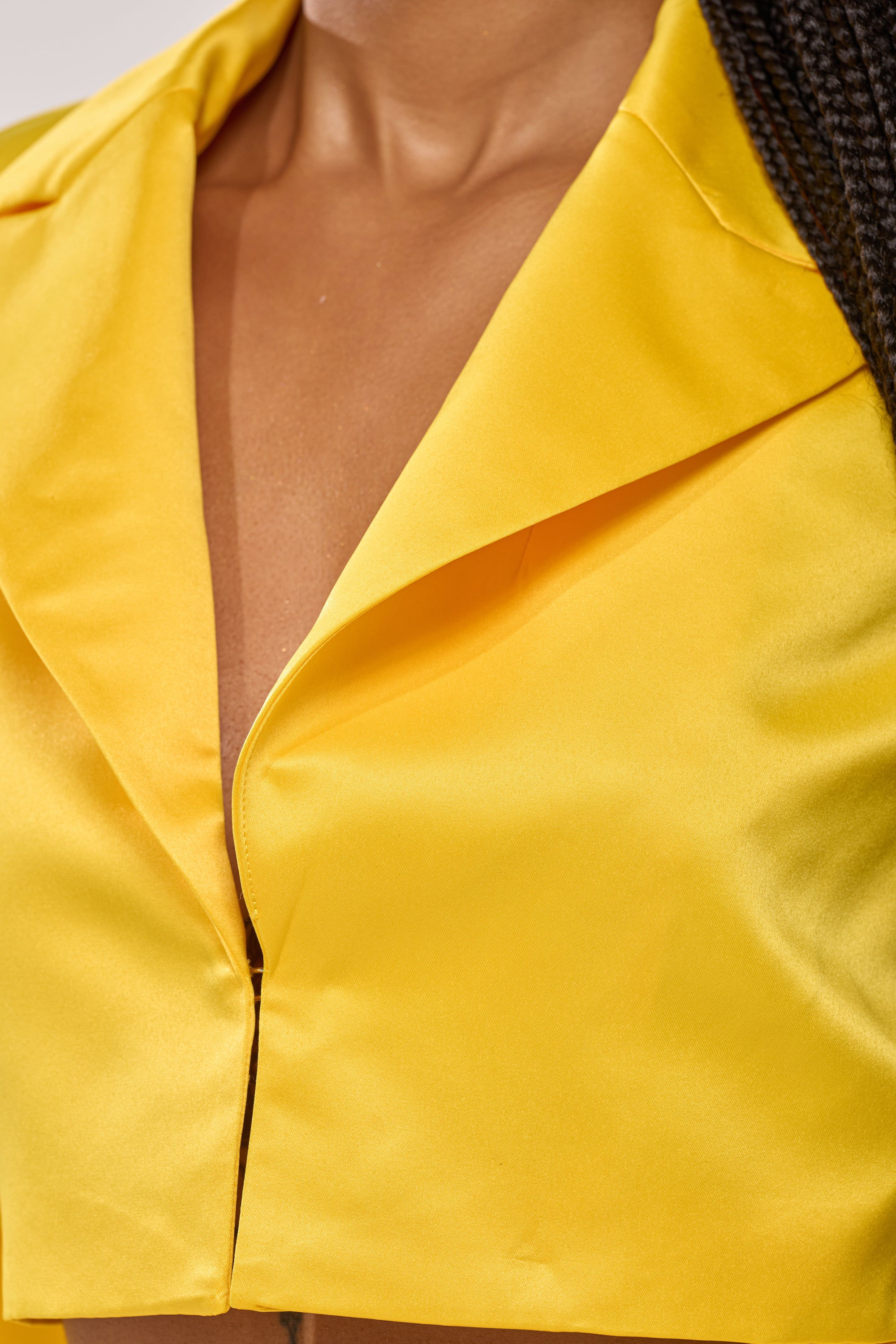 A close-up of a person wearing the BLOOMING CROP SATIN BLAZER in bright yellow, featuring a deep neckline and shiny satin that catches the light. Part of a braided hairstyle appears on the right side. 7
