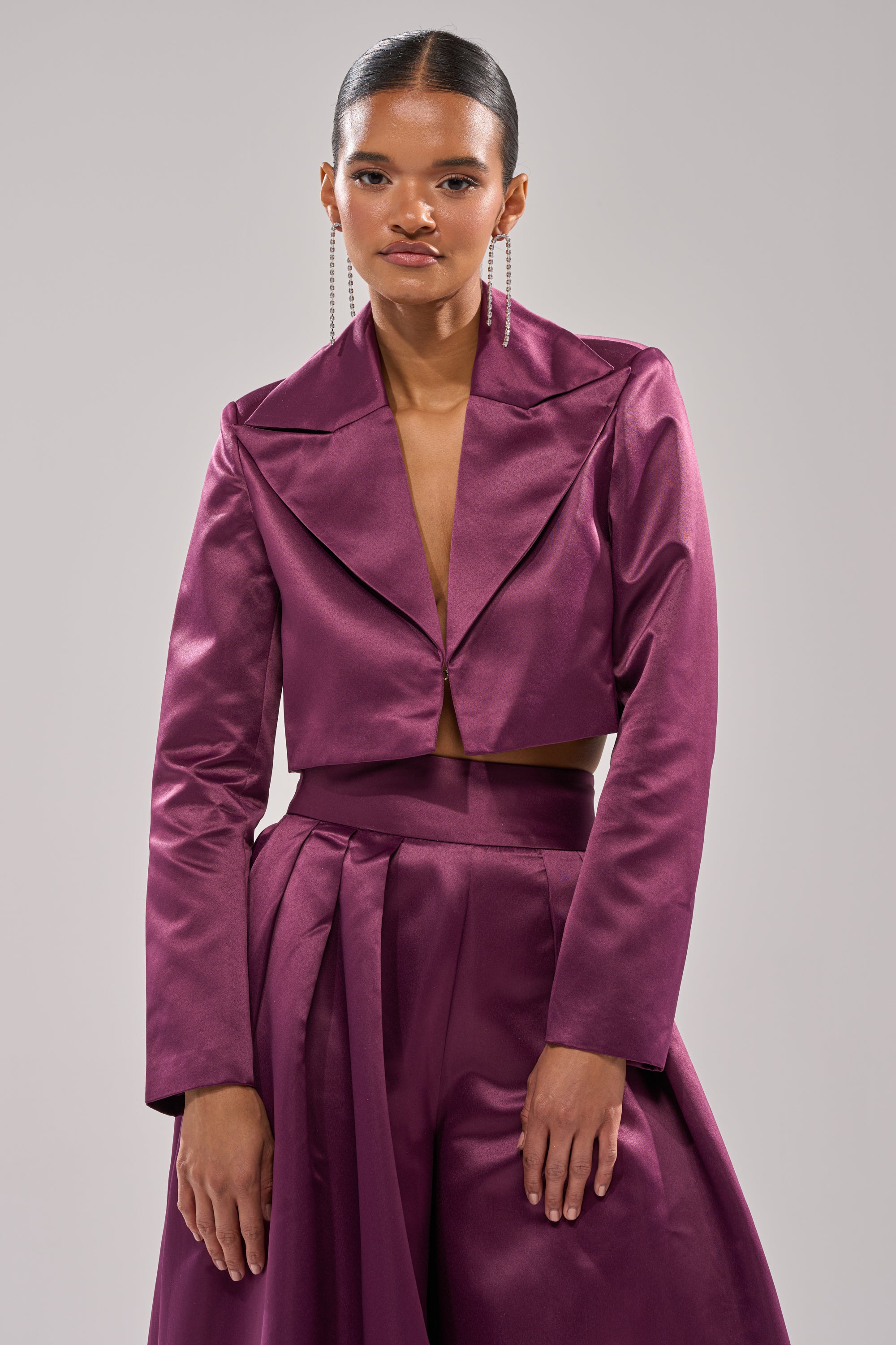 A woman wears the ON AND ON CROPPED SATIN BLAZER in shiny eggplant purple, paired with matching high-waisted pleated pants. Her sleek, pulled-back hair and long dangling earrings complete her chic look.