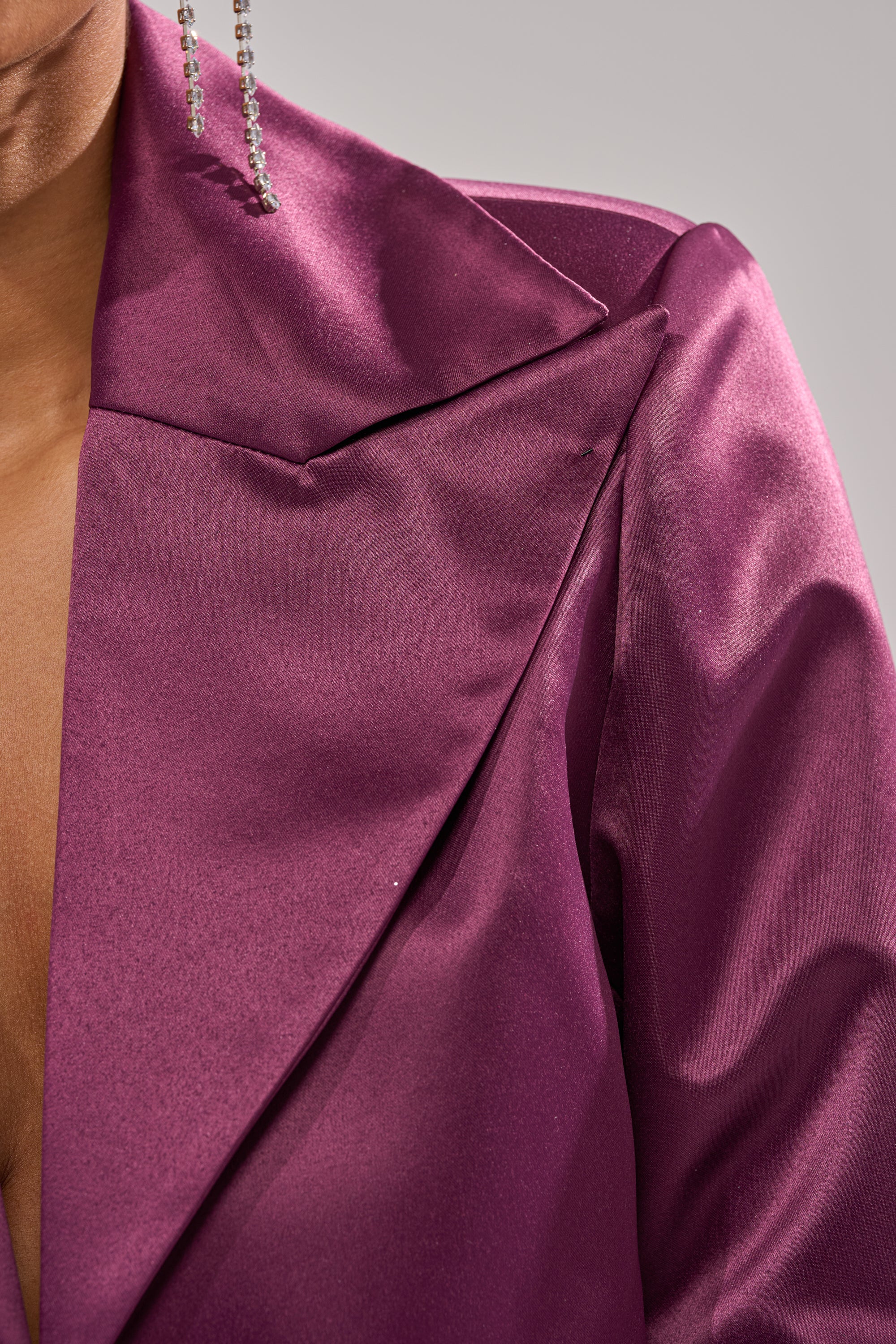 Close-up of the ON AND ON CROPPED SATIN BLAZER in glossy eggplant purple with wide lapels and a dangling beaded earring. The satin fabric shines against a neutral gray background, partially revealing the face.