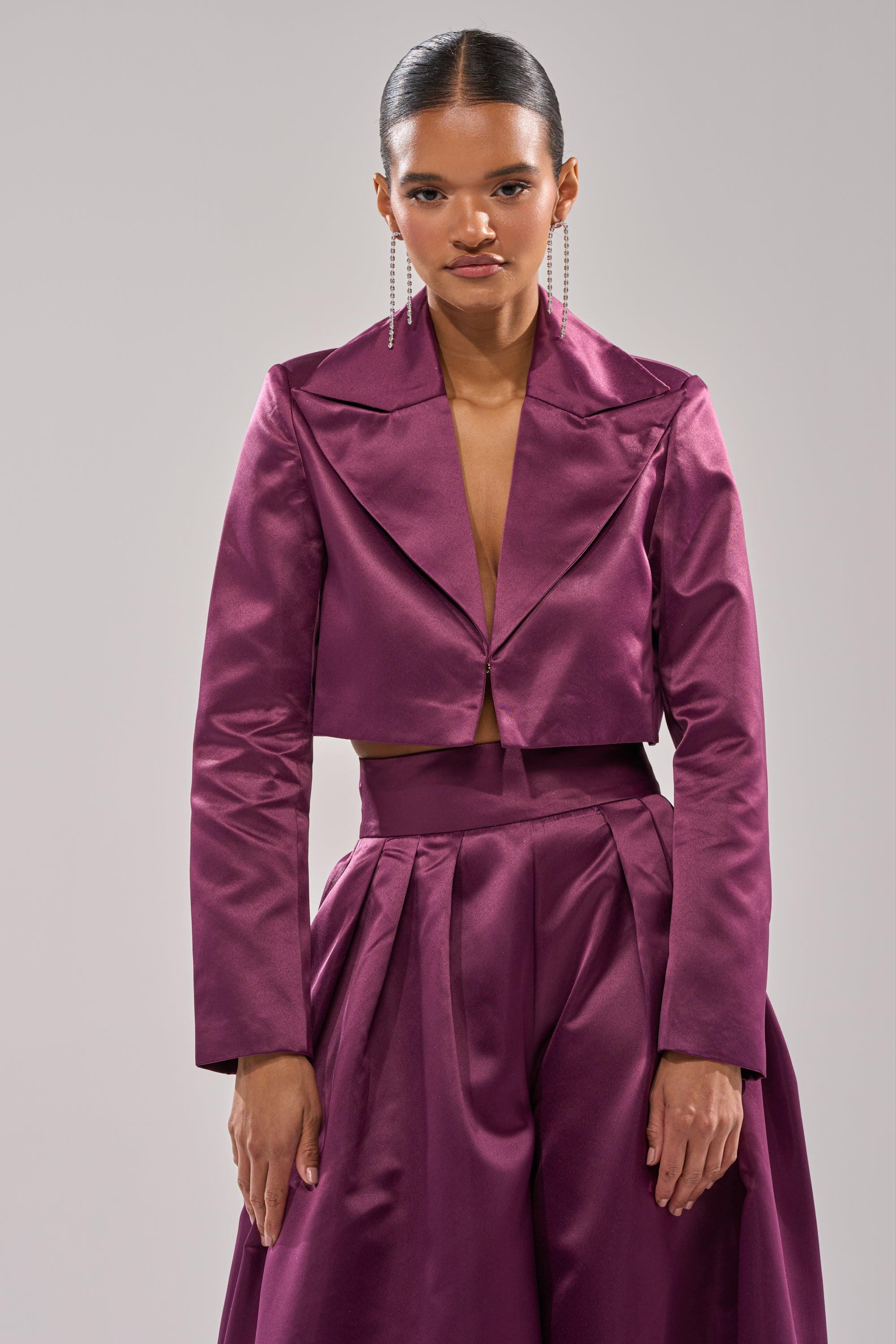 A woman models the ON AND ON CROPPED SATIN BLAZER in shiny eggplant purple with wide lapels and a matching high-waisted pleated skirt, paired with long drop earrings and slicked-back hair against a plain background.