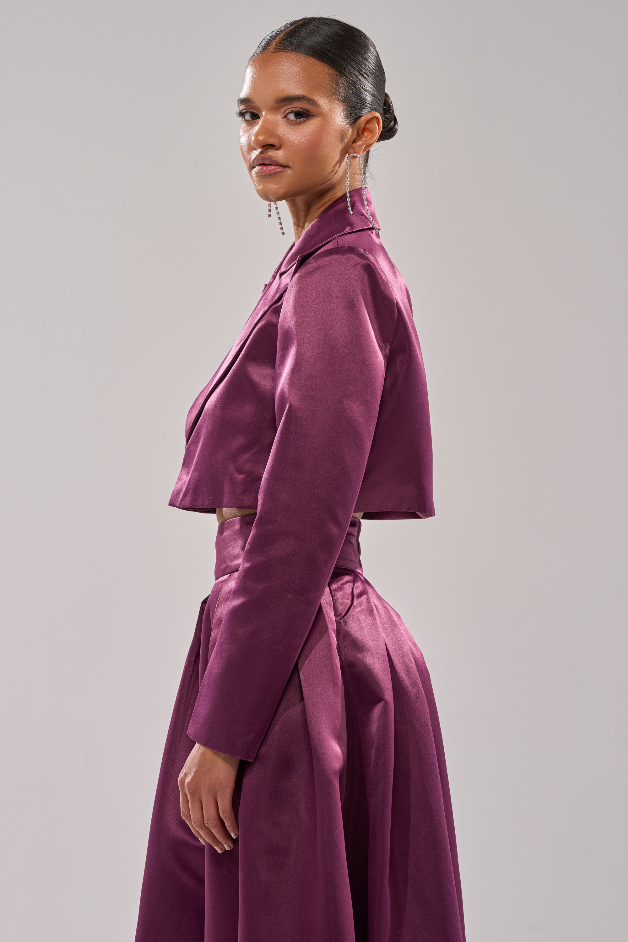 A woman stands sideways against a plain background, wearing the ON AND ON CROPPED SATIN BLAZER in deep eggplant purple with a matching long skirt. Her sleek bun and dangling earrings complete the look.