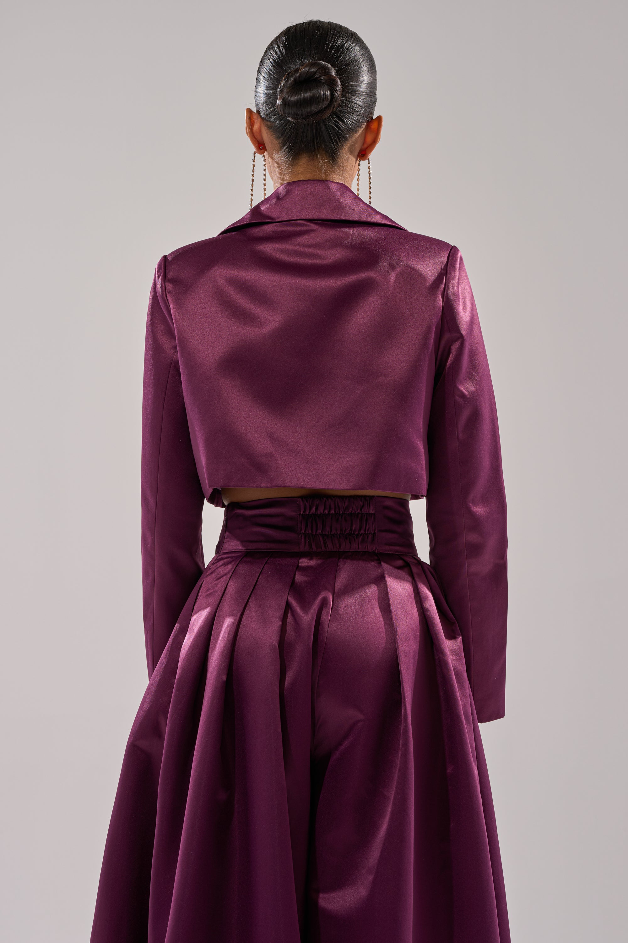 A person faces away, wearing the ON AND ON CROPPED SATIN BLAZER in eggplant purple with a matching high-waisted pleated skirt, hair in a sleek bun and long earrings. The background is plain and light-colored.